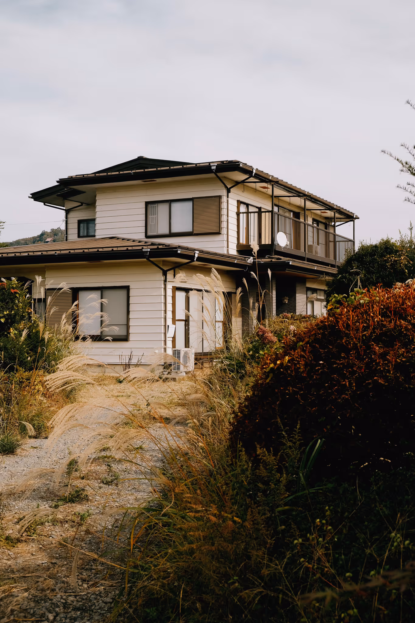 Two-story house with white siding, brown window shutters, and a balcony surrounded by tall grasses and shrubs.