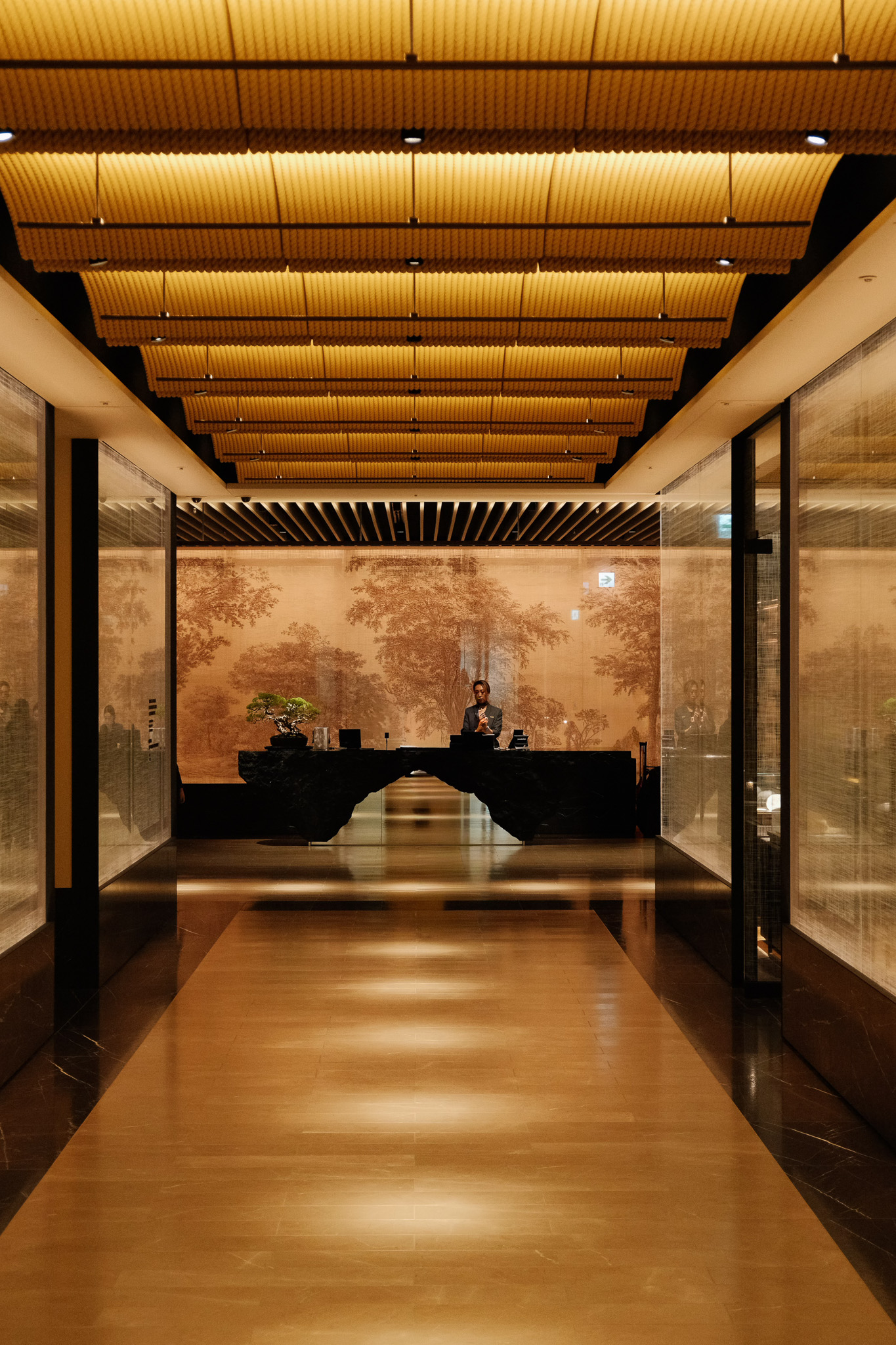 Modern hotel lobby with warm wooden floor, unique ceiling lighting, and a person standing behind a black reception desk.