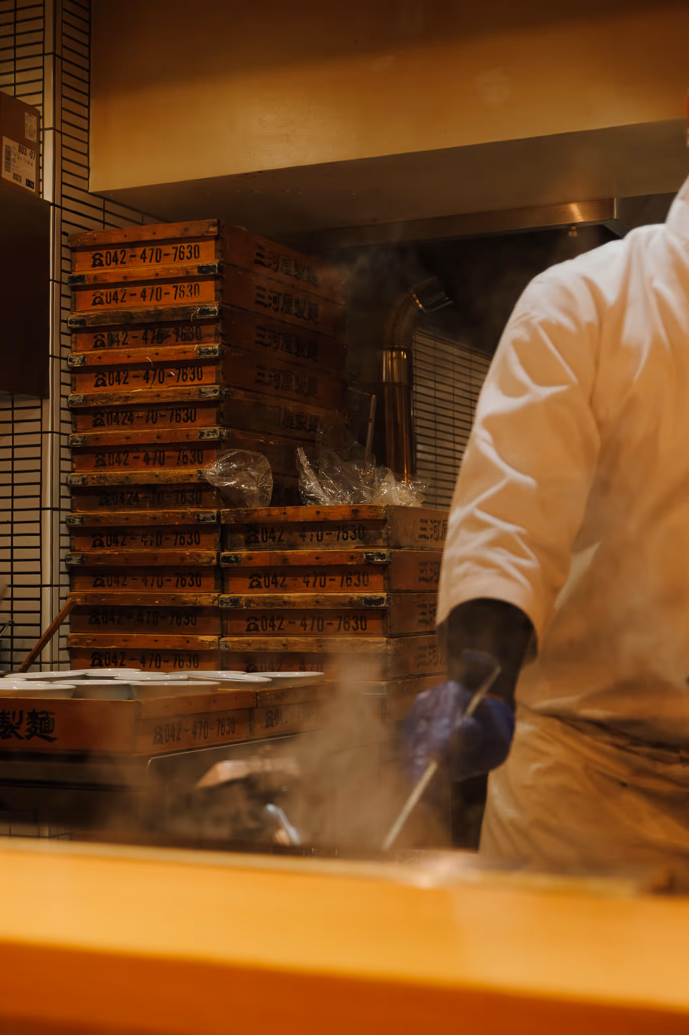 Chef in white uniform cooking in a kitchen with stacks of wooden trays and steam rising.