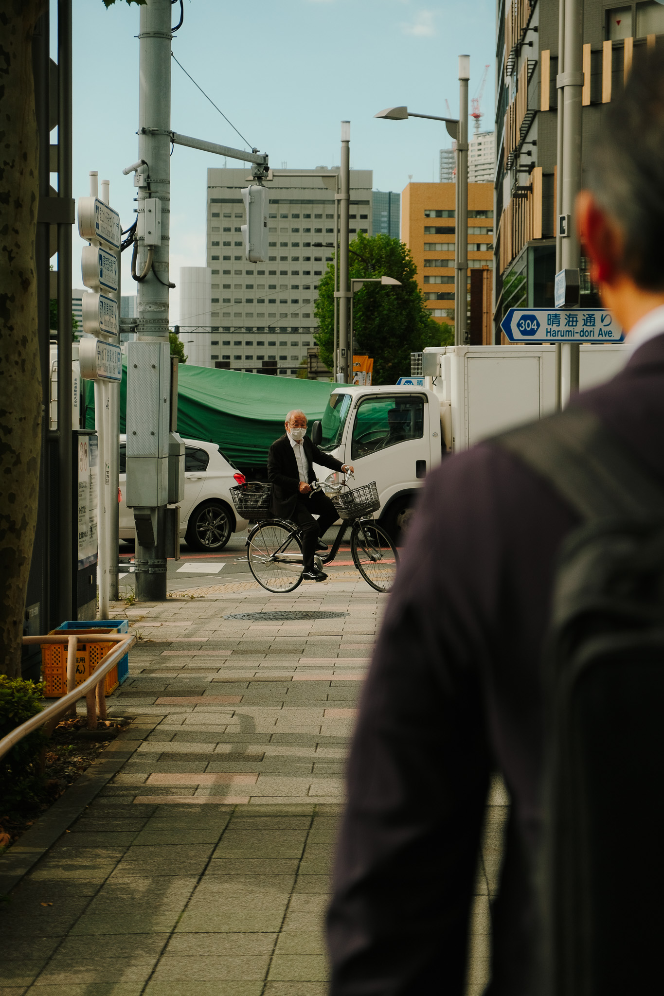 Older man wearing a mask riding a bicycle on a city sidewalk with buildings and vehicles in the background.