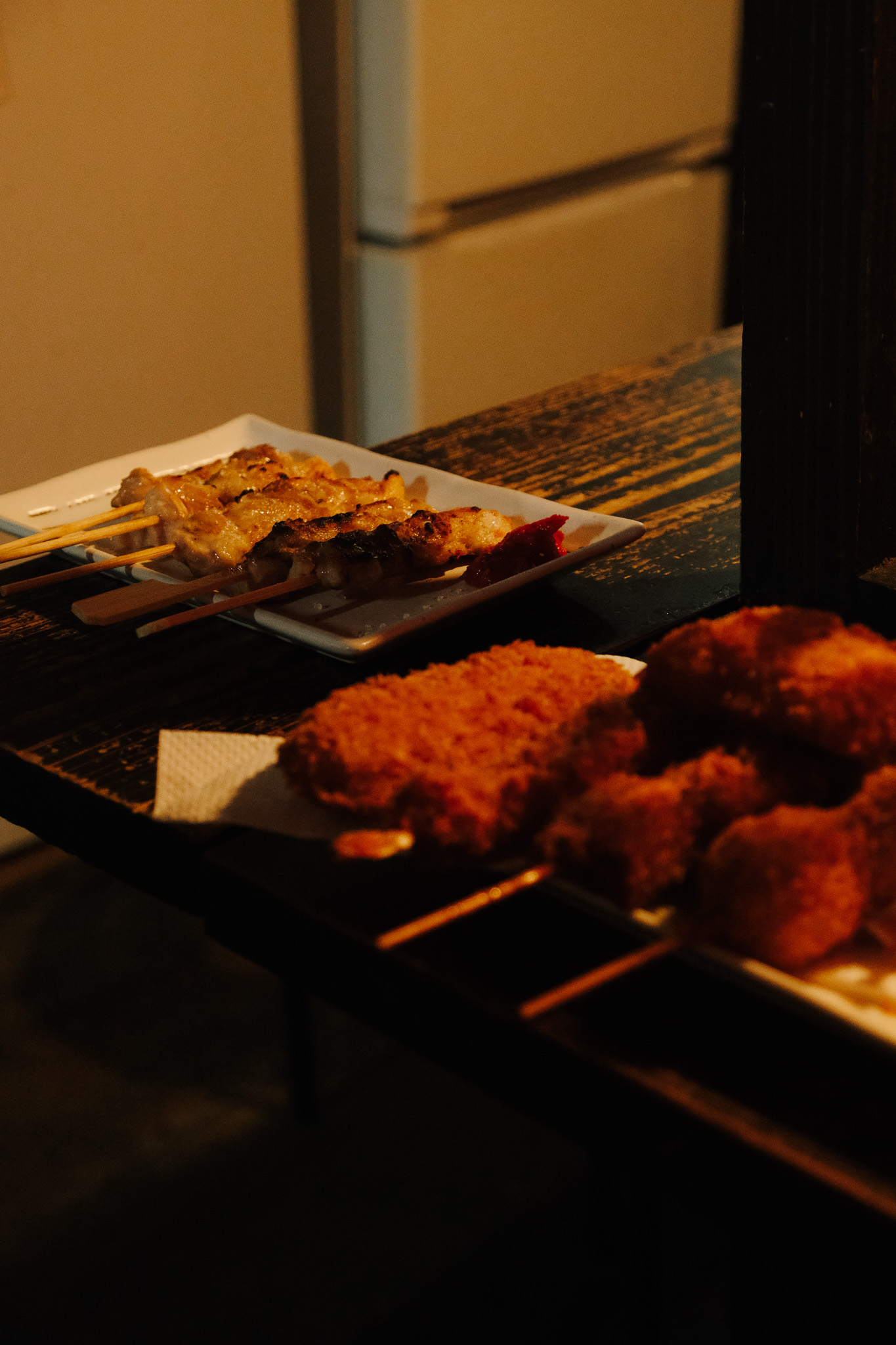 Grilled meat skewers and breaded fried food on white plates on a dark wooden table.
