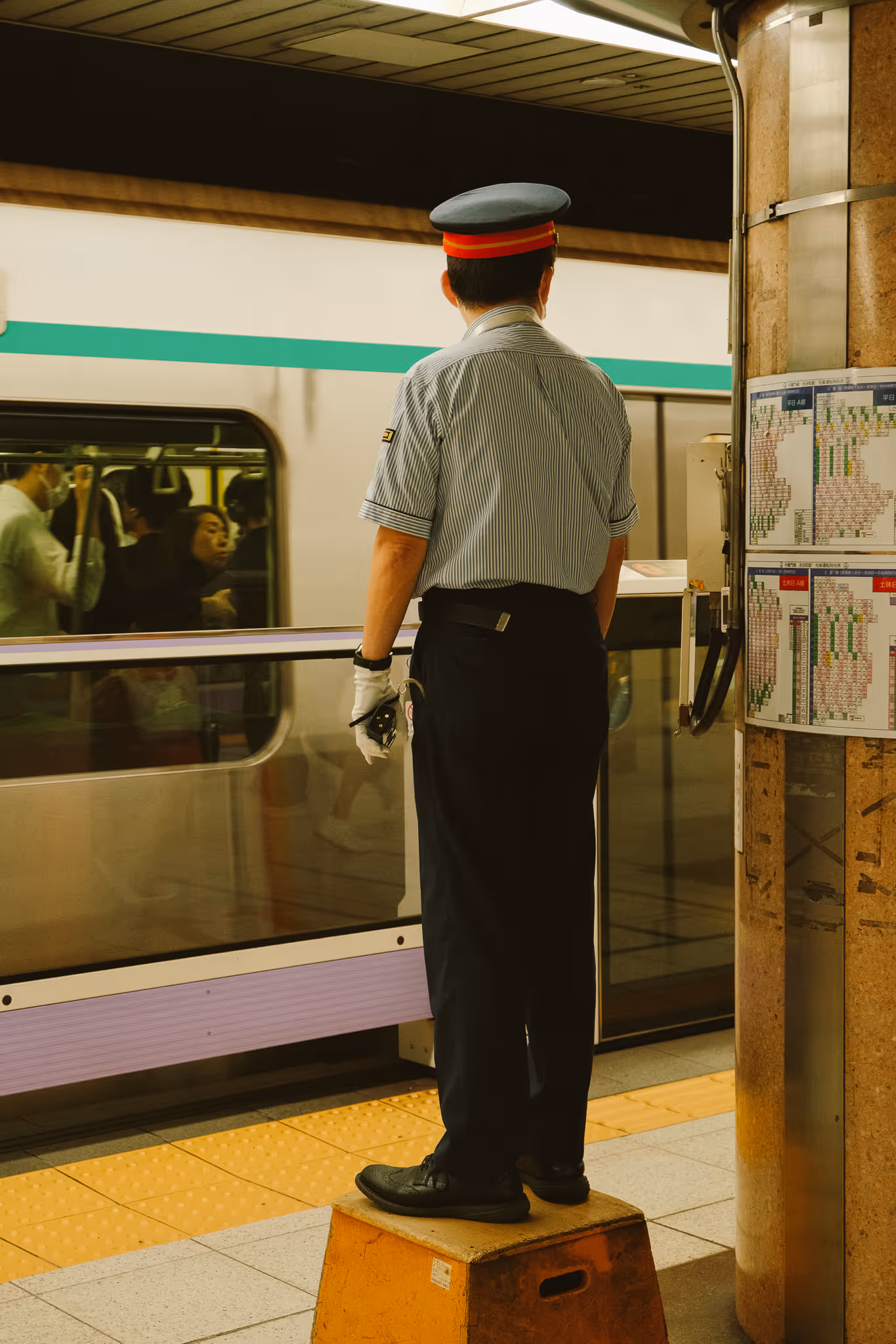 Train station worker in uniform standing on a wooden box facing a passing train with passengers inside.