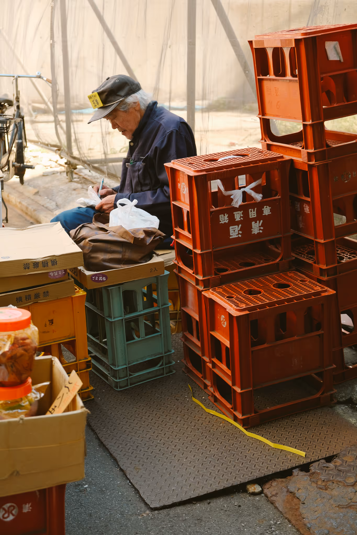Elderly man wearing a cap, sitting among plastic crates and boxes, writing on a piece of paper.