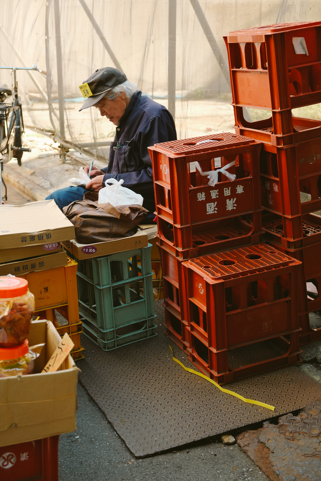 Elderly man wearing a cap, sitting among plastic crates and boxes, writing on a piece of paper.