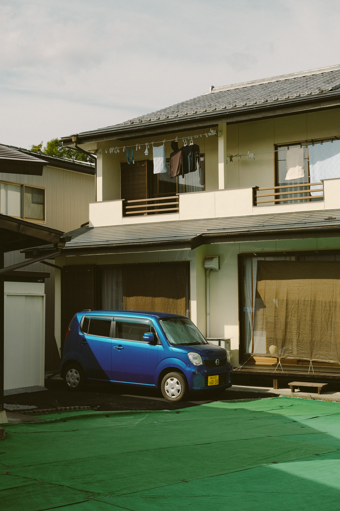 Blue compact car parked in front of a beige two-story house with laundry hanging on a clothesline on the upper balcony.