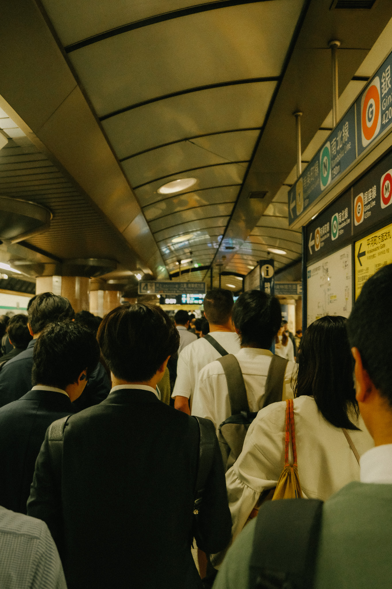 Crowded subway station corridor with commuters seen from behind under a curved ceiling with directional signs in Japanese.