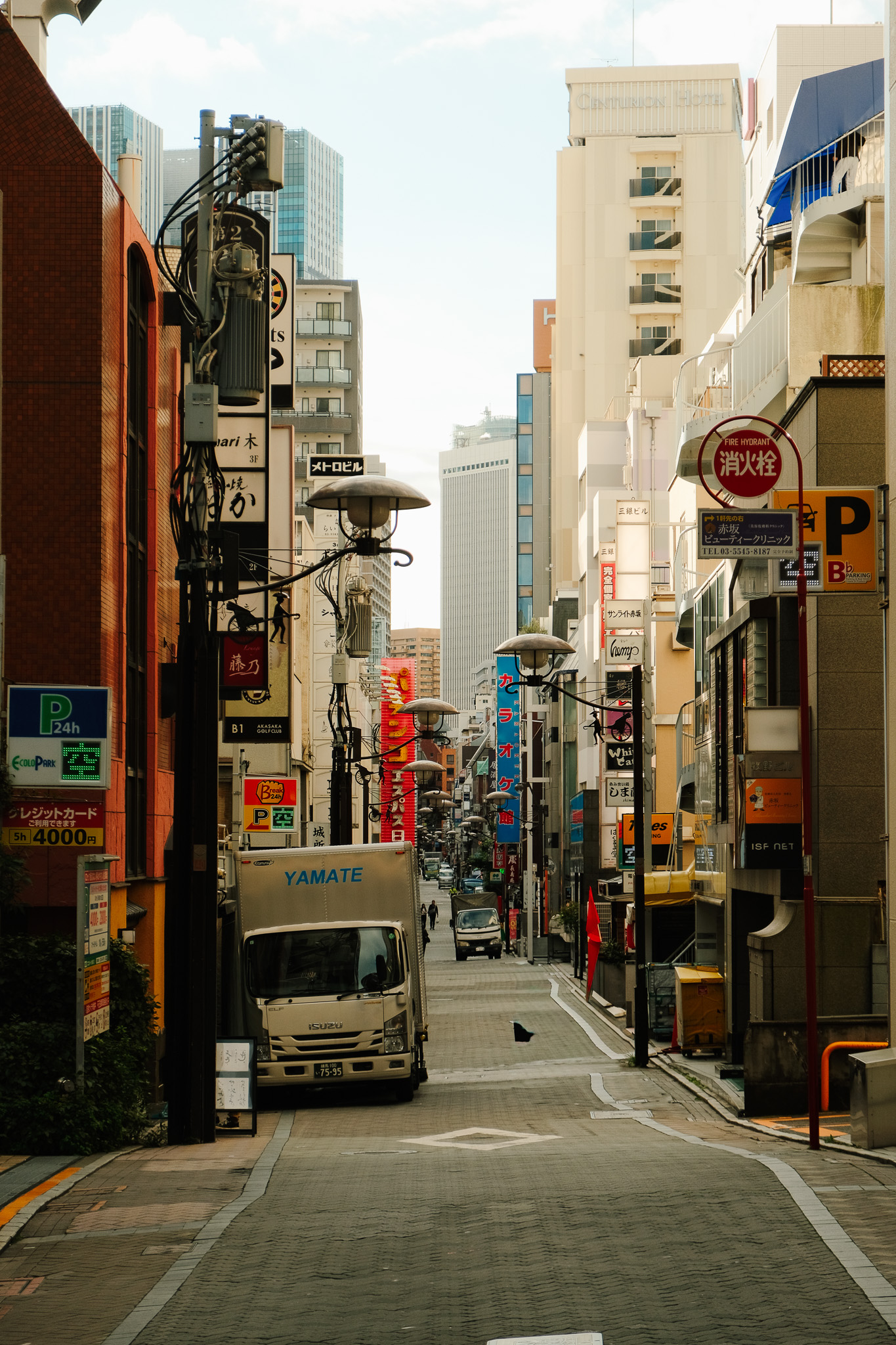 Narrow urban street in Japan lined with buildings featuring many signs in Japanese, a parked delivery truck, street lamps, and a few pedestrians in the distance.