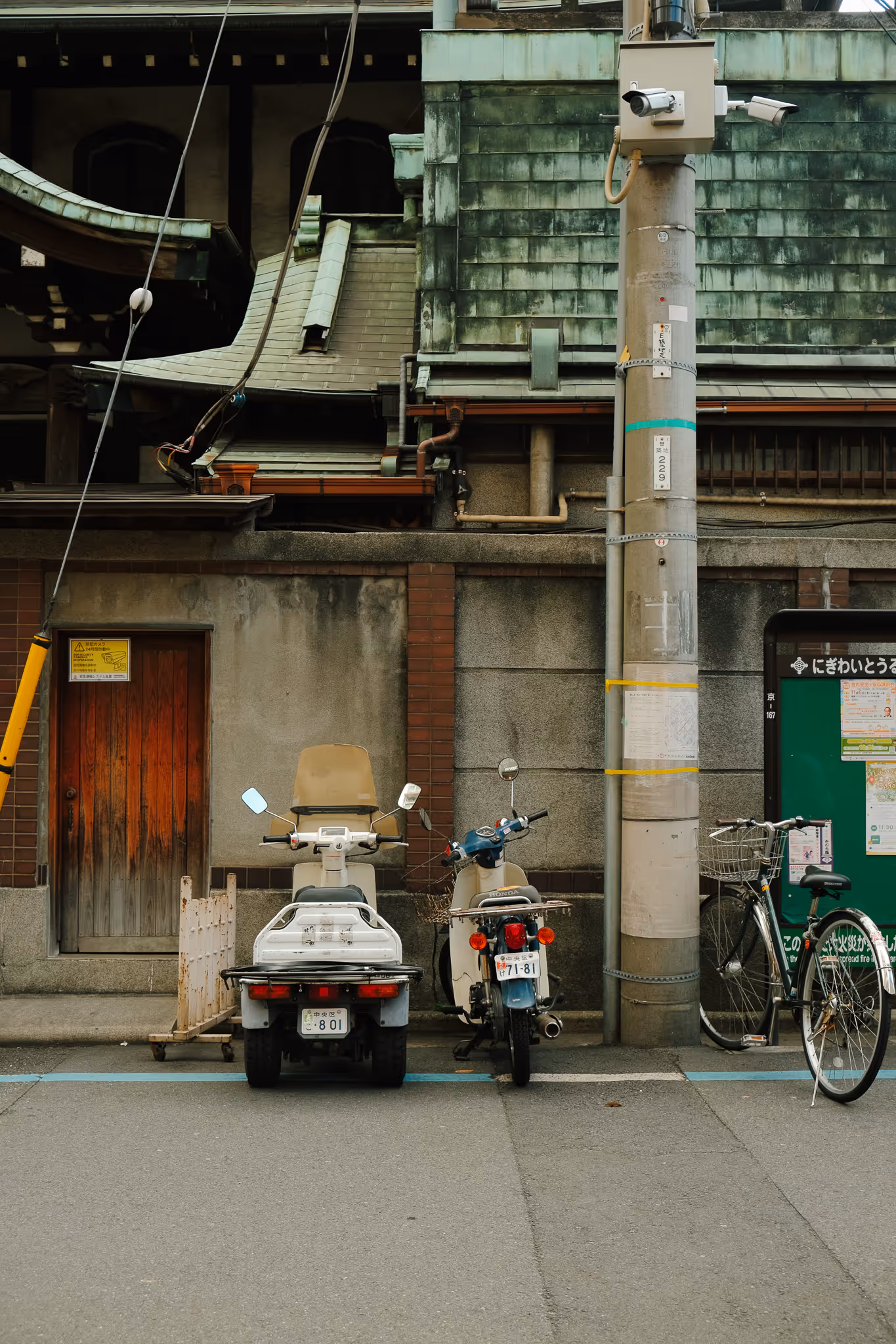 Two motor scooters and a bicycle parked on the street in front of a weathered building with a wooden door and green tiled roof in a quiet urban setting.