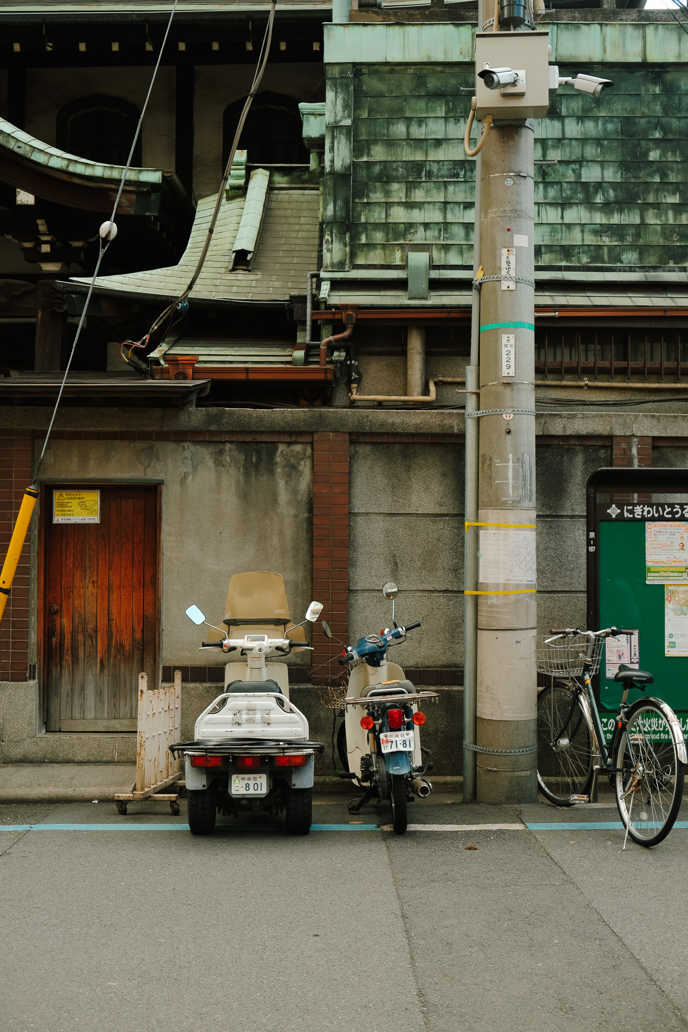 Two motor scooters and a bicycle parked on the street in front of a weathered building with a wooden door and green tiled roof in a quiet urban setting.