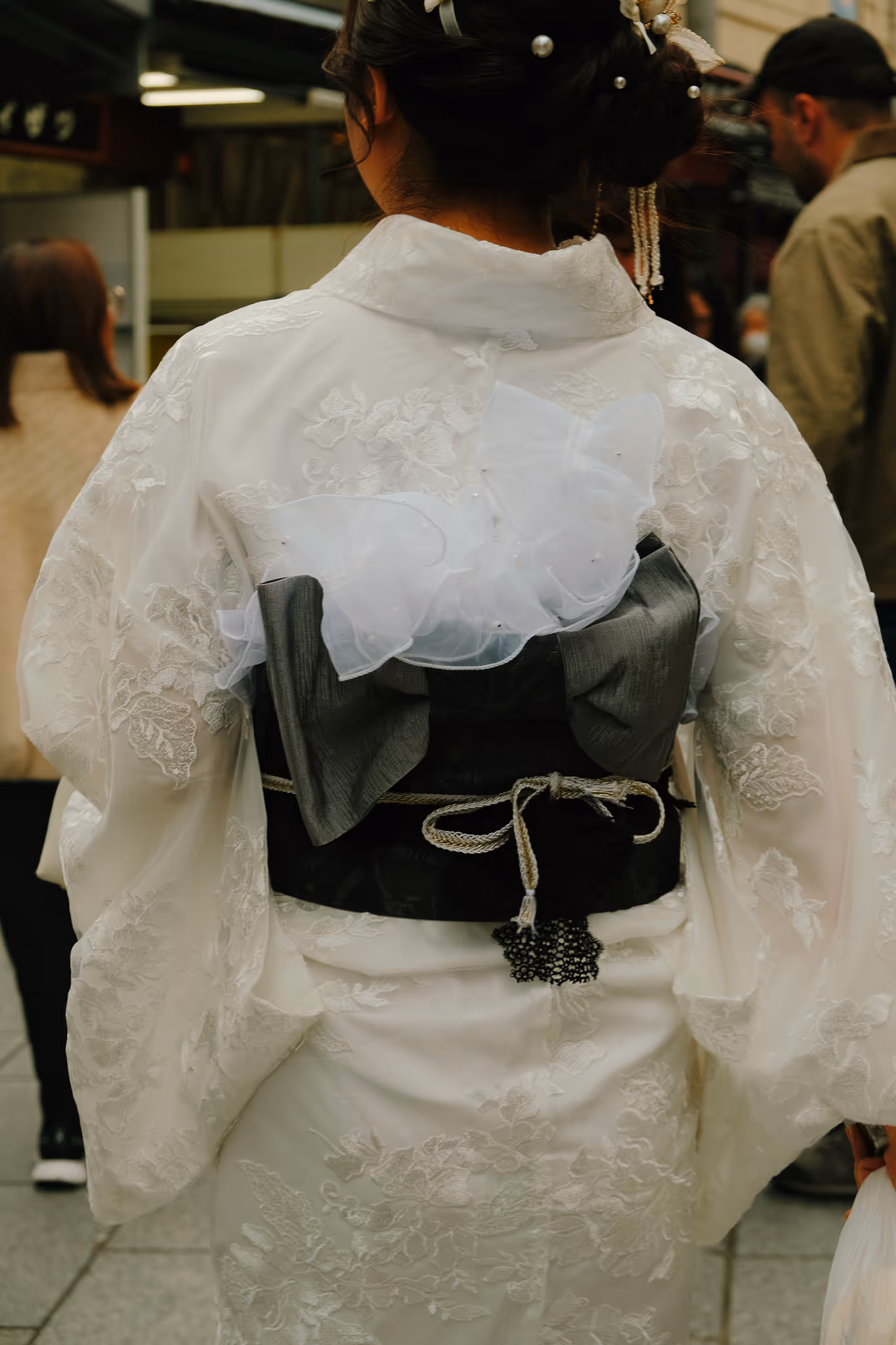 Back view of a woman wearing a white lace kimono with a black obi belt adorned with a large white bow and decorative accessories.
