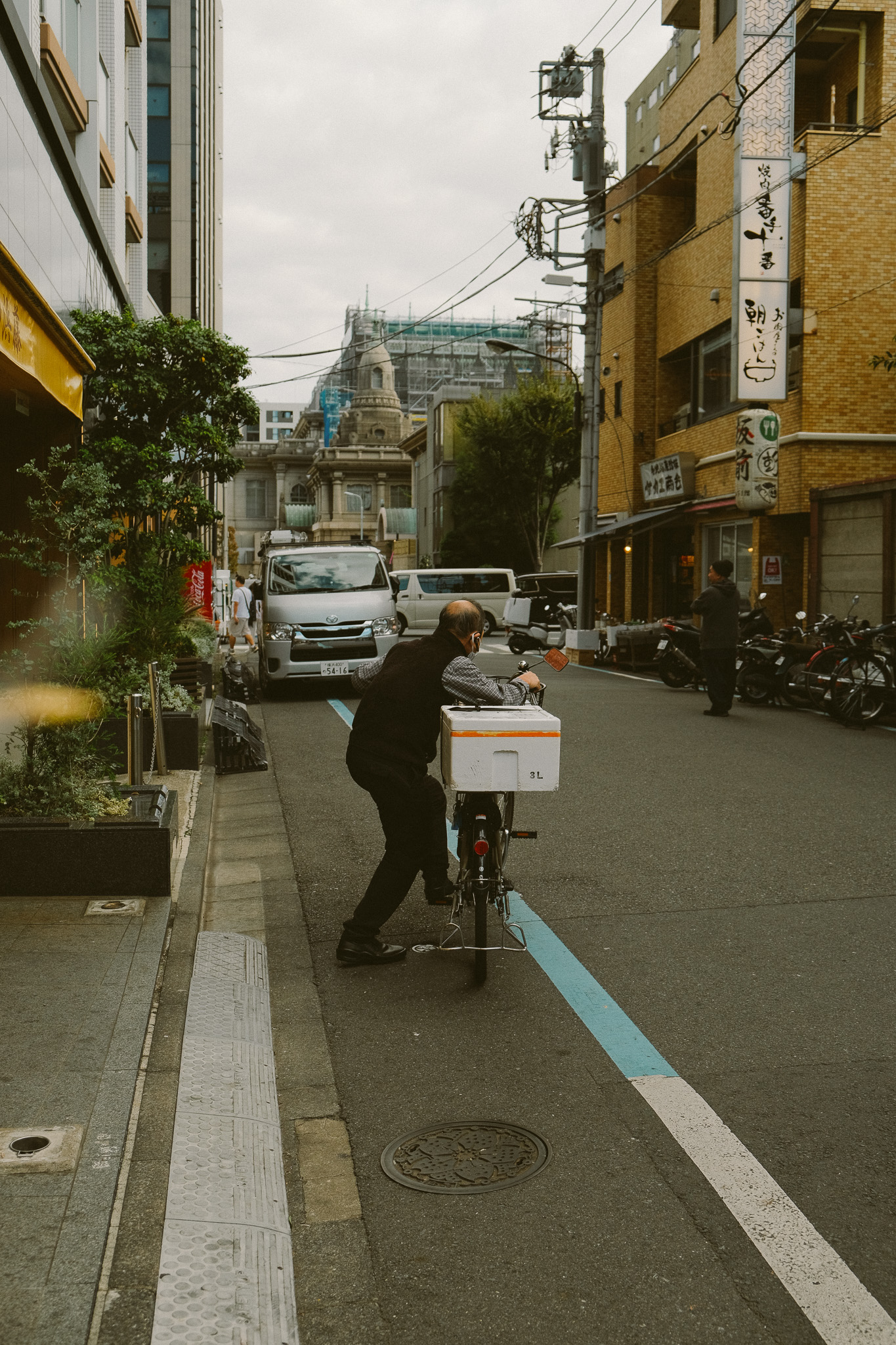 Man pushing a bicycle with a white box on a narrow city street lined with parked cars, bicycles, and buildings.