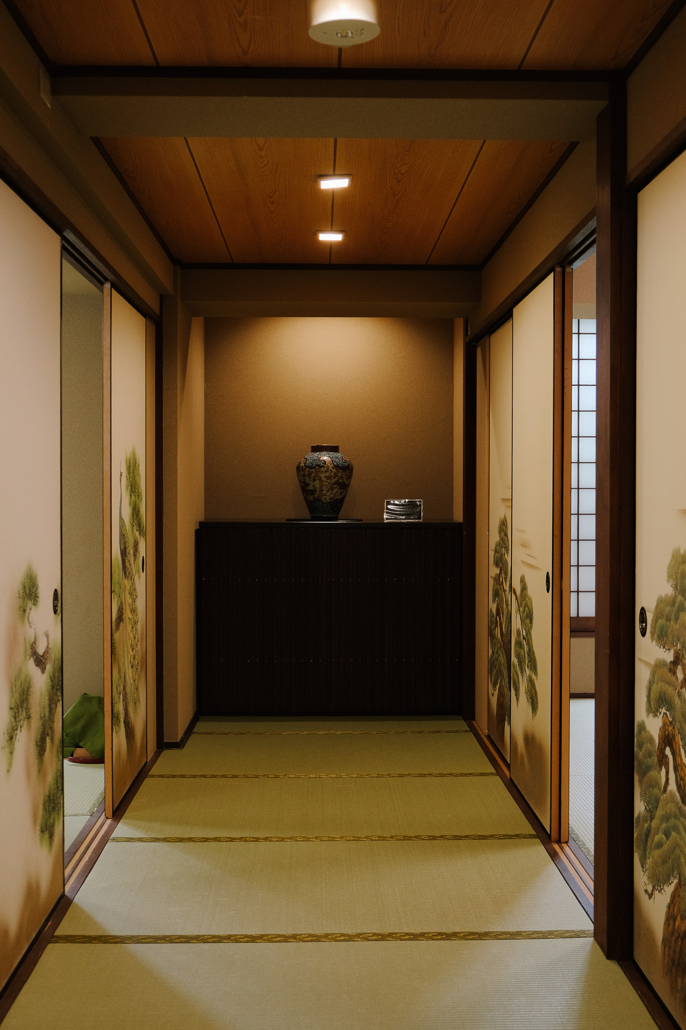 Traditional Japanese room corridor with sliding doors featuring tree paintings and a vase on a cabinet at the end.