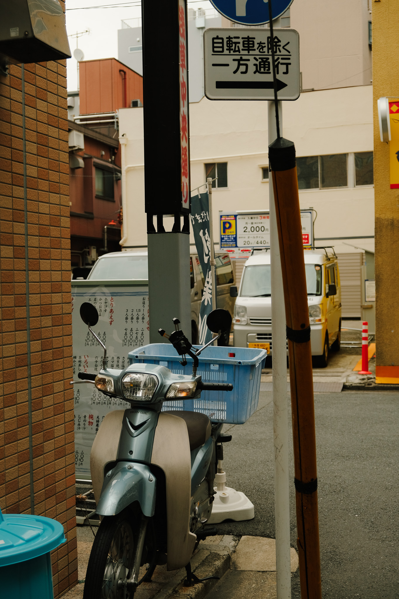 Blue scooter with a blue plastic basket parked on a sidewalk between a brick wall and a pole, with cars and Japanese signs in the background.