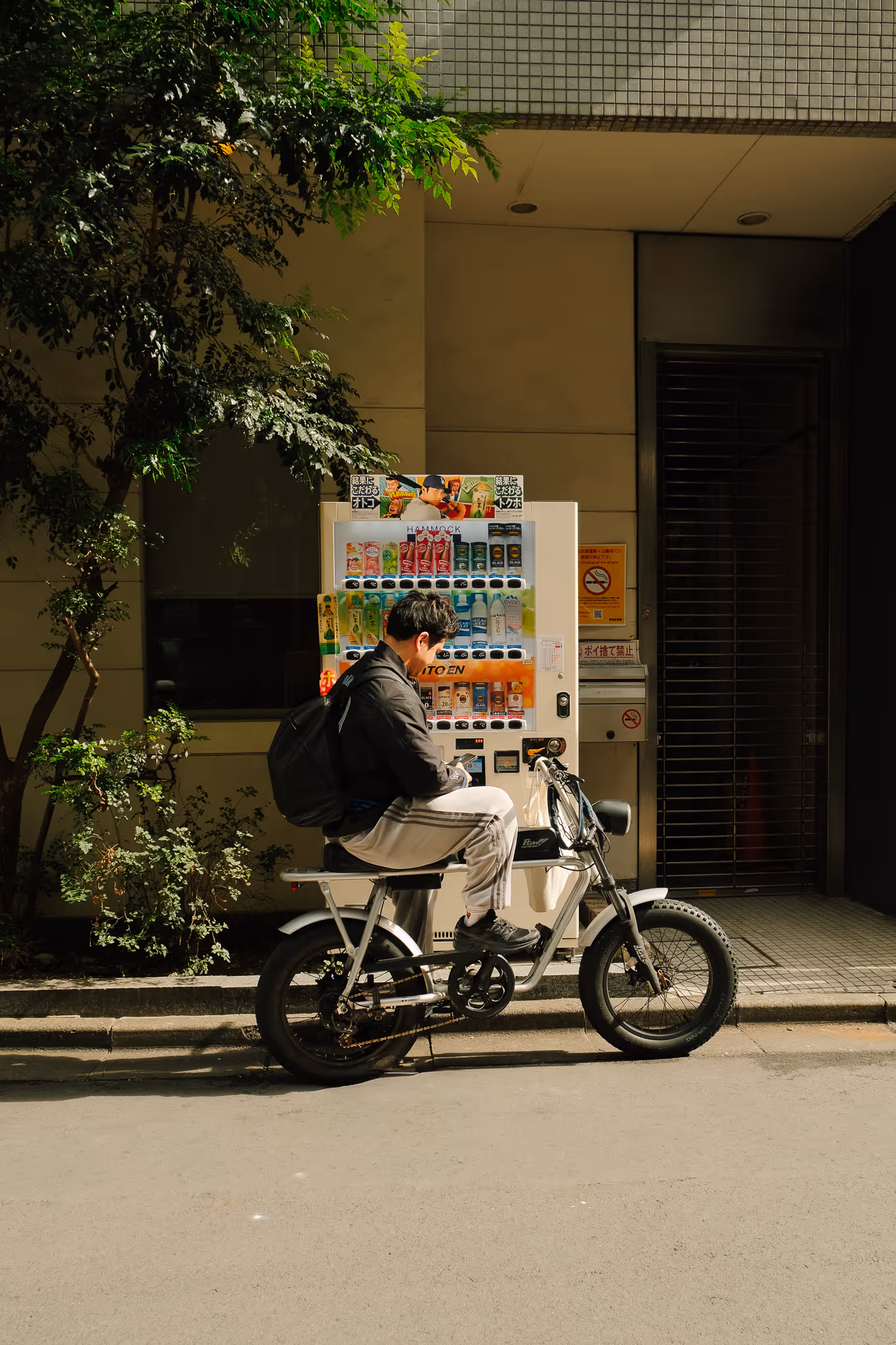 Man wearing a black jacket and gray pants sitting on a motorbike in front of a vending machine on a sunlit street.
