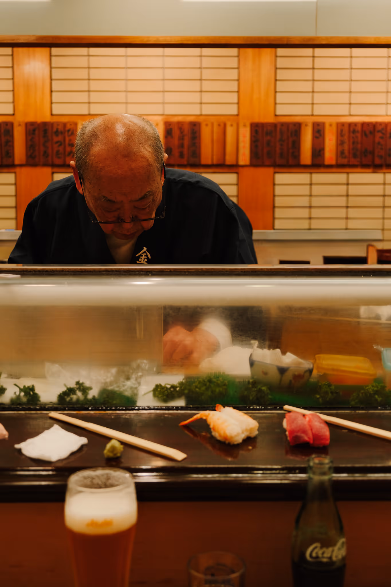 Sushi chef preparing food behind glass counter with pieces of sushi, chopsticks, and beverages in foreground.