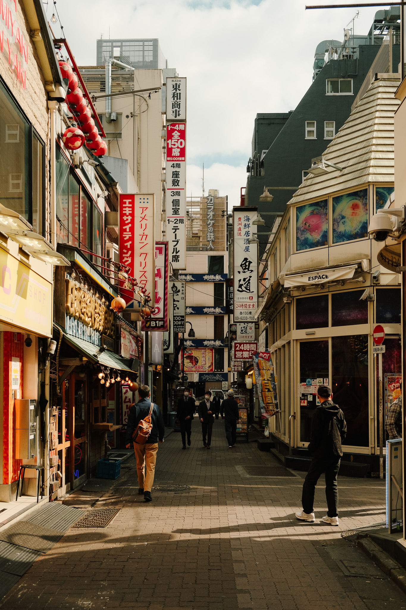 Narrow urban street lined with shops and illuminated signs in Japanese, with pedestrians walking and standing under a partly cloudy sky.