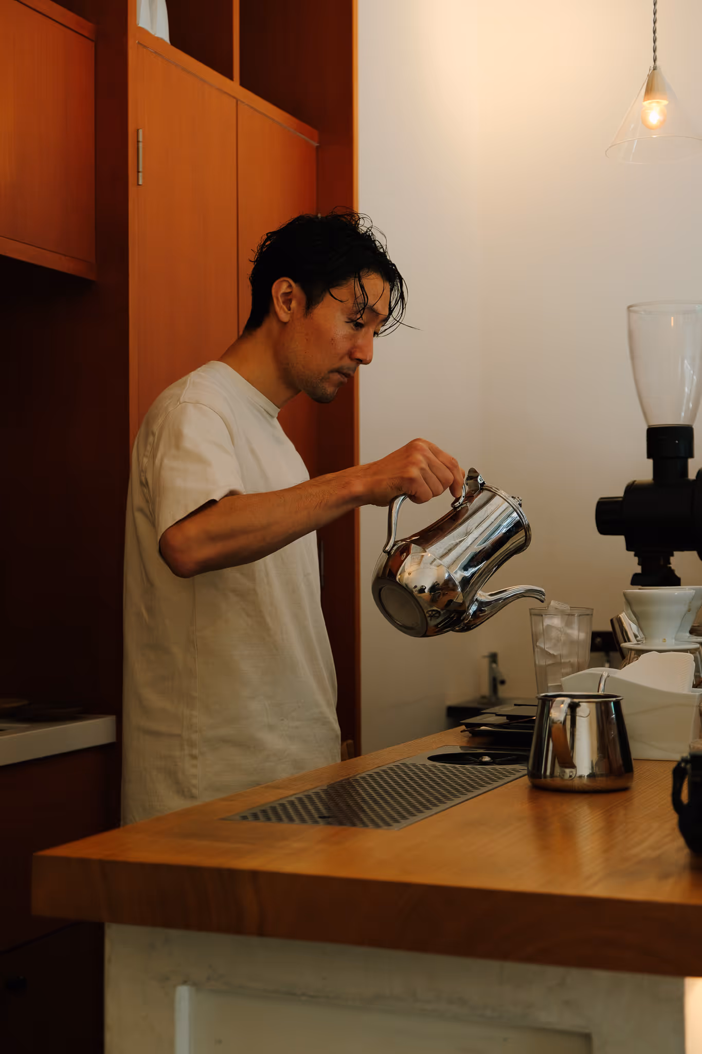 Man pouring water from a shiny metal kettle behind a wooden counter in a coffee shop.
