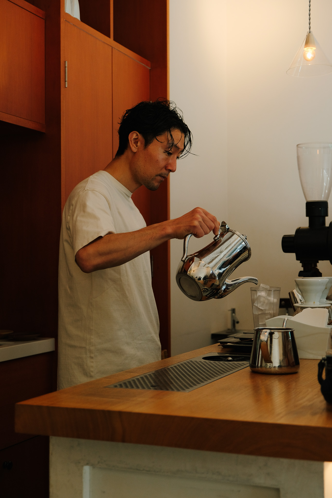 Man pouring water from a shiny metal kettle behind a wooden counter in a coffee shop.