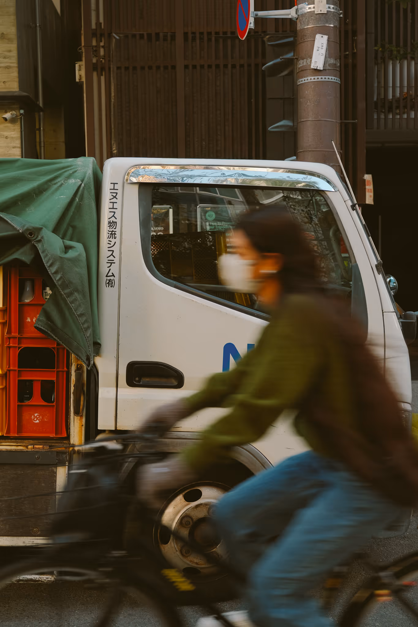 Blurred person wearing a mask riding a bicycle past a white truck with Japanese text on its door and red crates covered with a green tarp.
