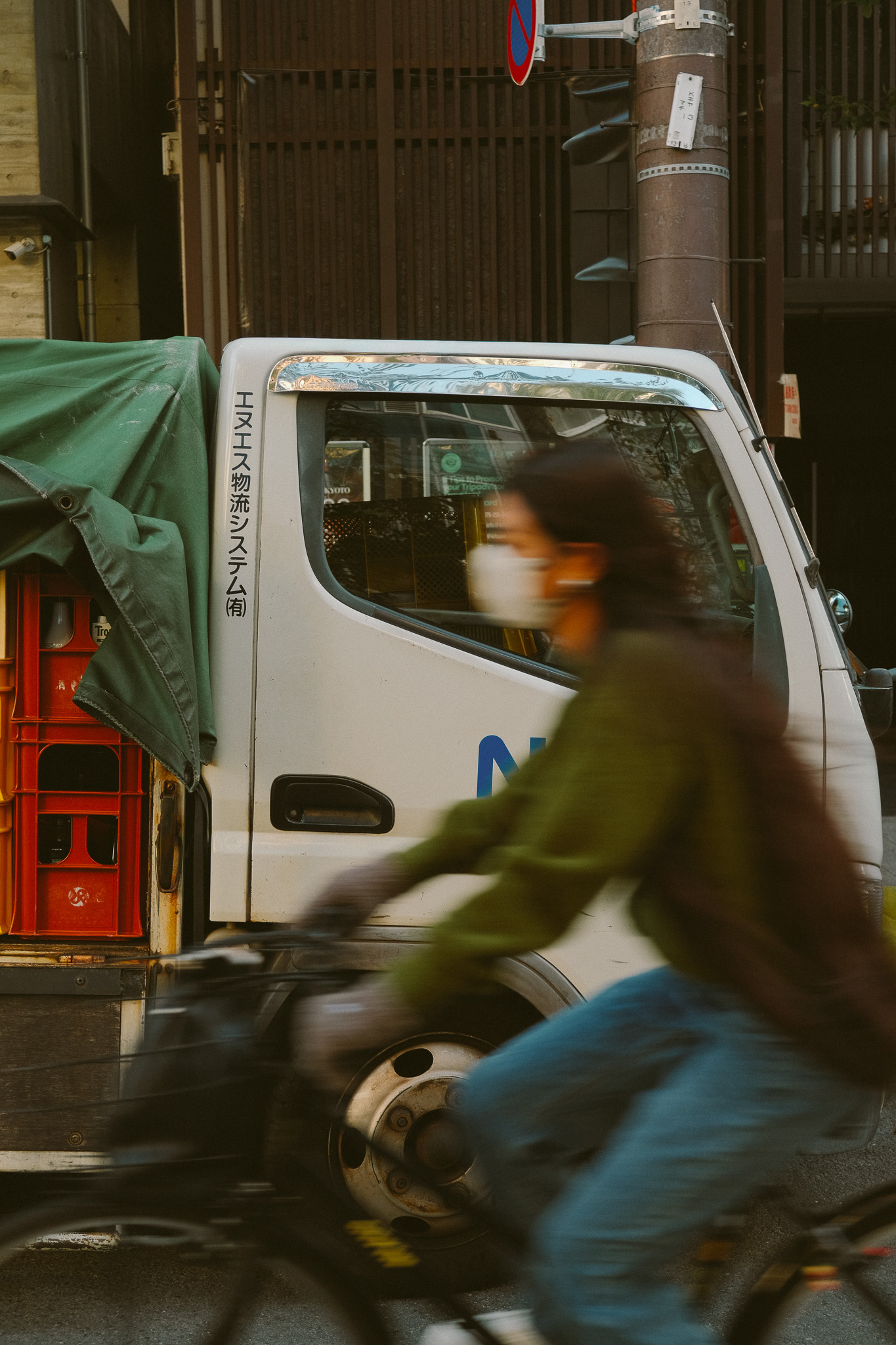 Blurred person wearing a mask riding a bicycle past a white truck with Japanese text on its door and red crates covered with a green tarp.