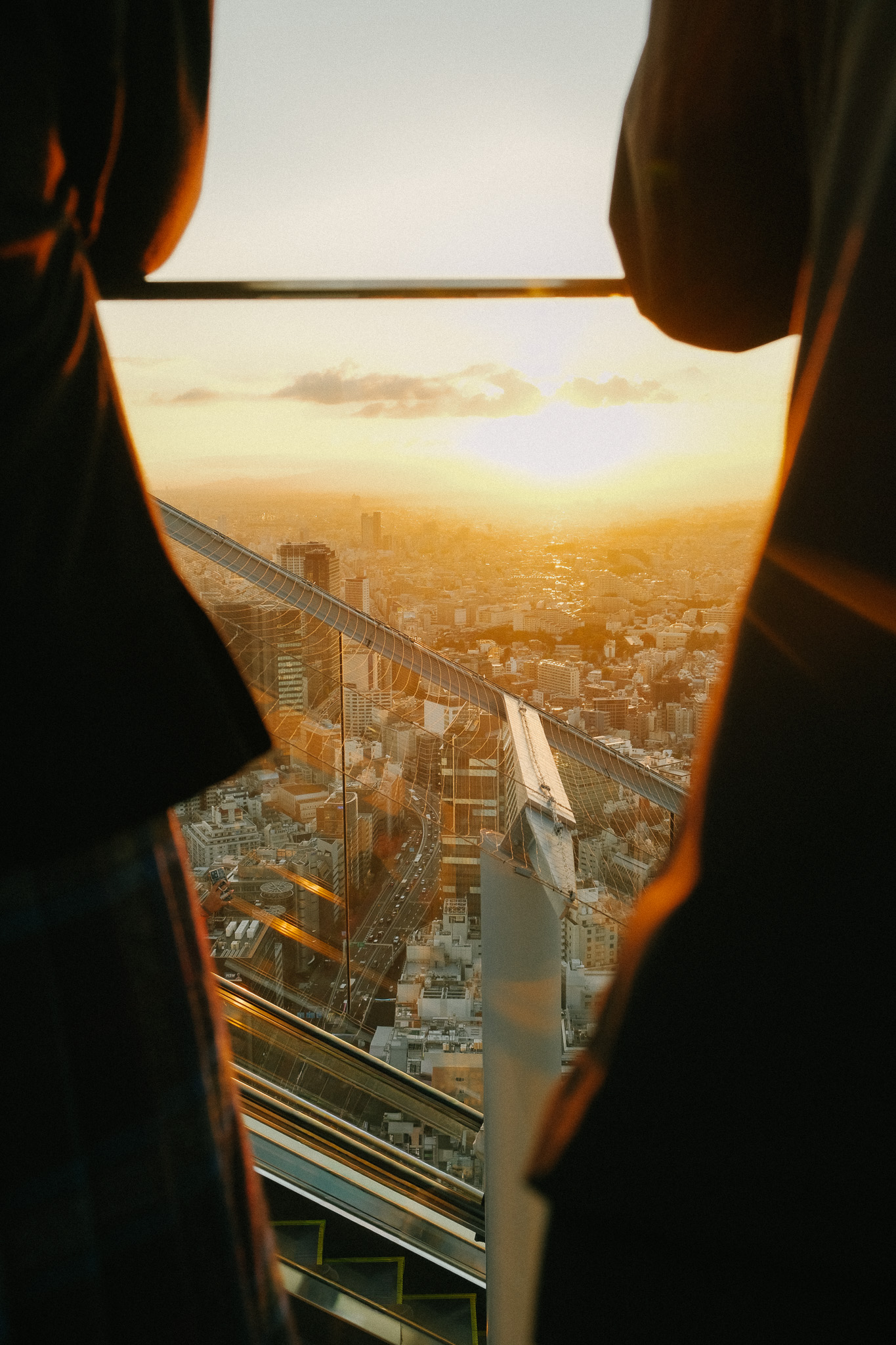 Sunset view over a cityscape seen through a glass railing with parts of two people silhouetted in the foreground.