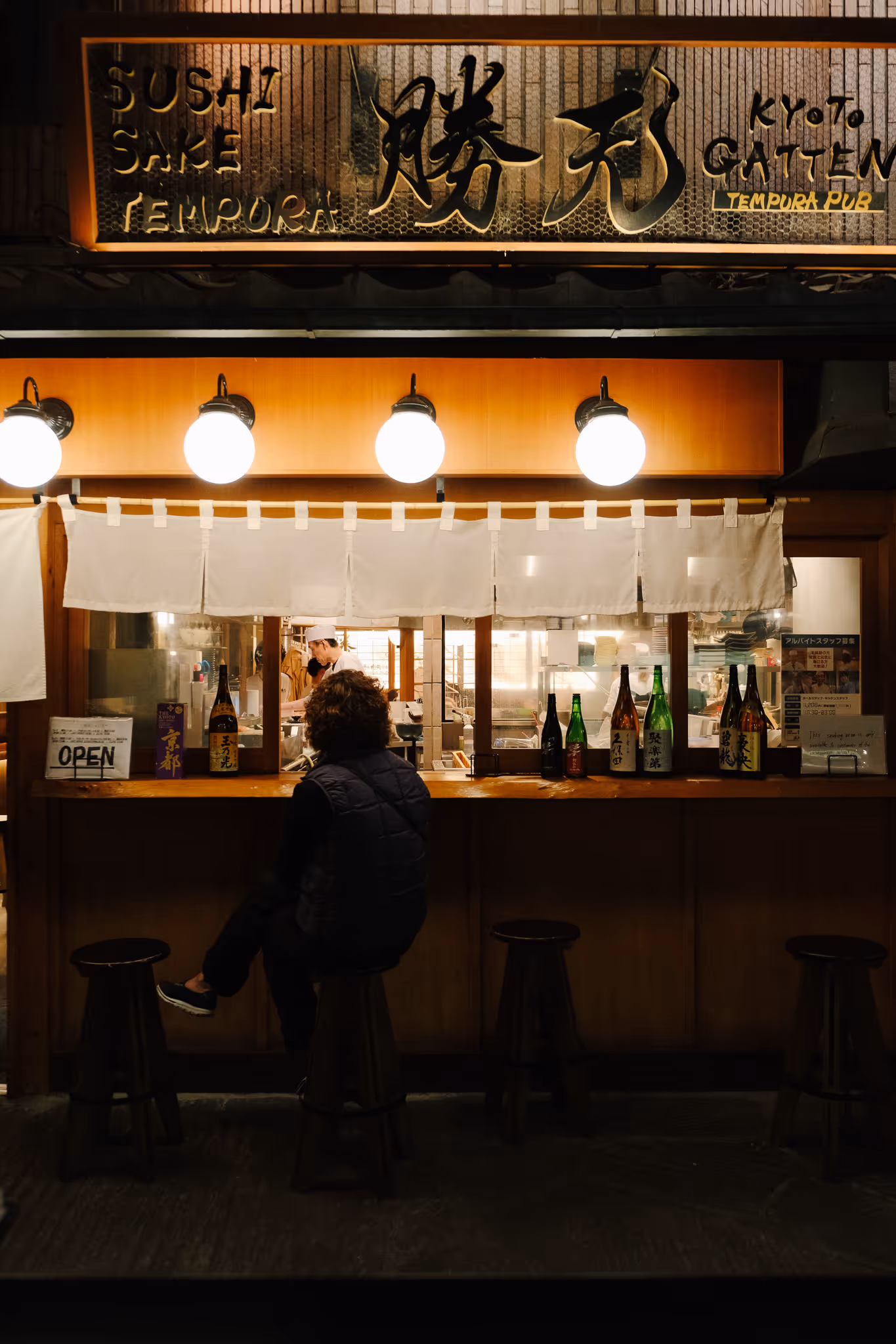 A person sitting on a stool at a wooden counter of a small illuminated Japanese tempura pub with bottles and a chef working inside.