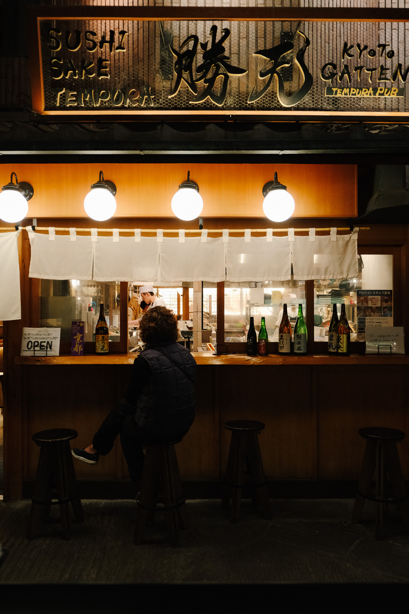 A person sitting on a stool at a wooden counter of a small illuminated Japanese tempura pub with bottles and a chef working inside.