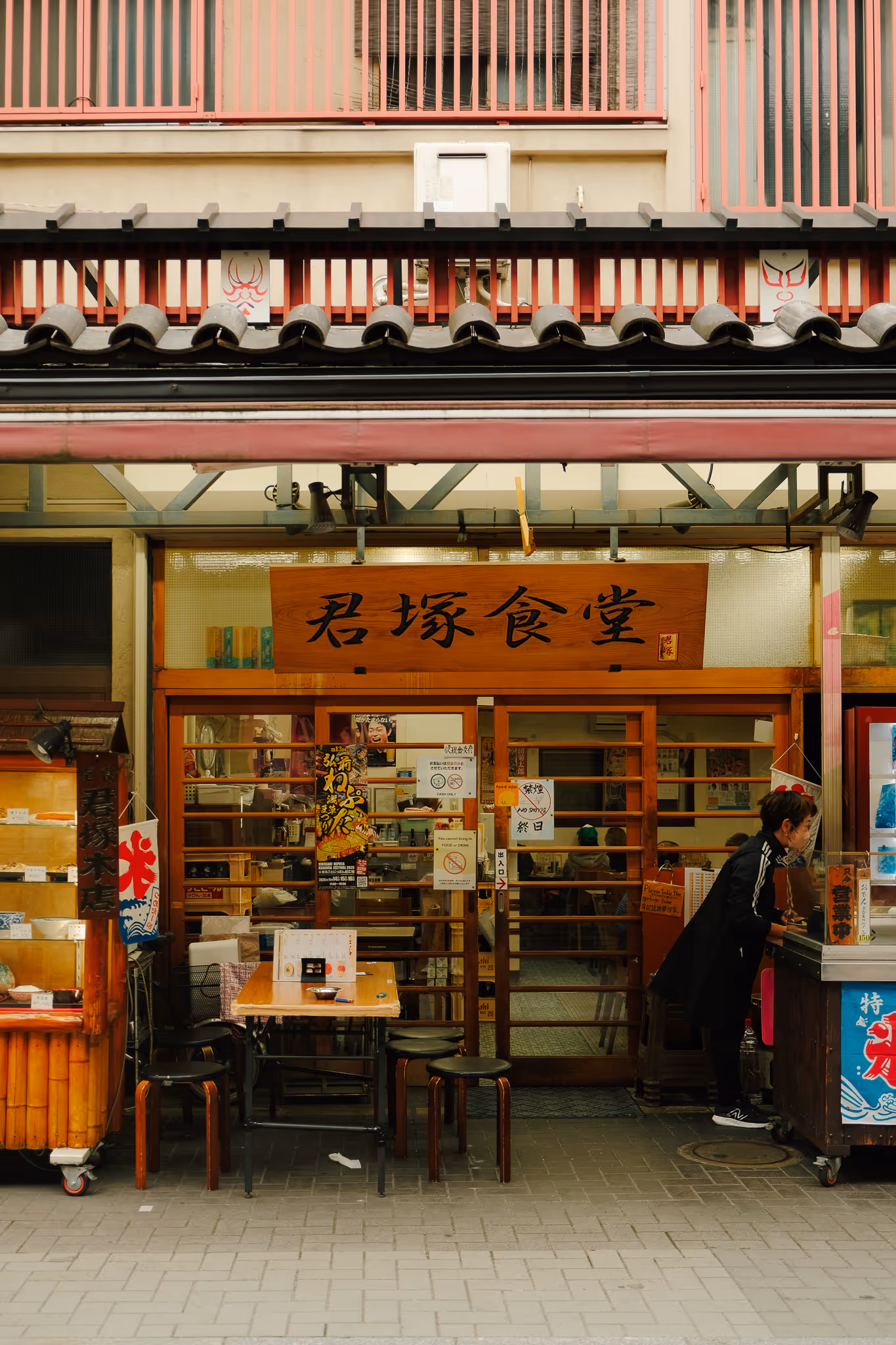 Exterior of a Japanese restaurant with wooden stools and a table outside, a woman leaning over the counter, and a sign with Japanese characters above the entrance.
