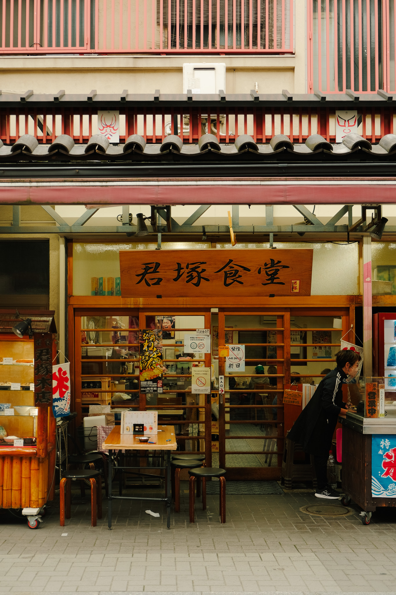 Exterior of a Japanese restaurant with wooden stools and a table outside, a woman leaning over the counter, and a sign with Japanese characters above the entrance.