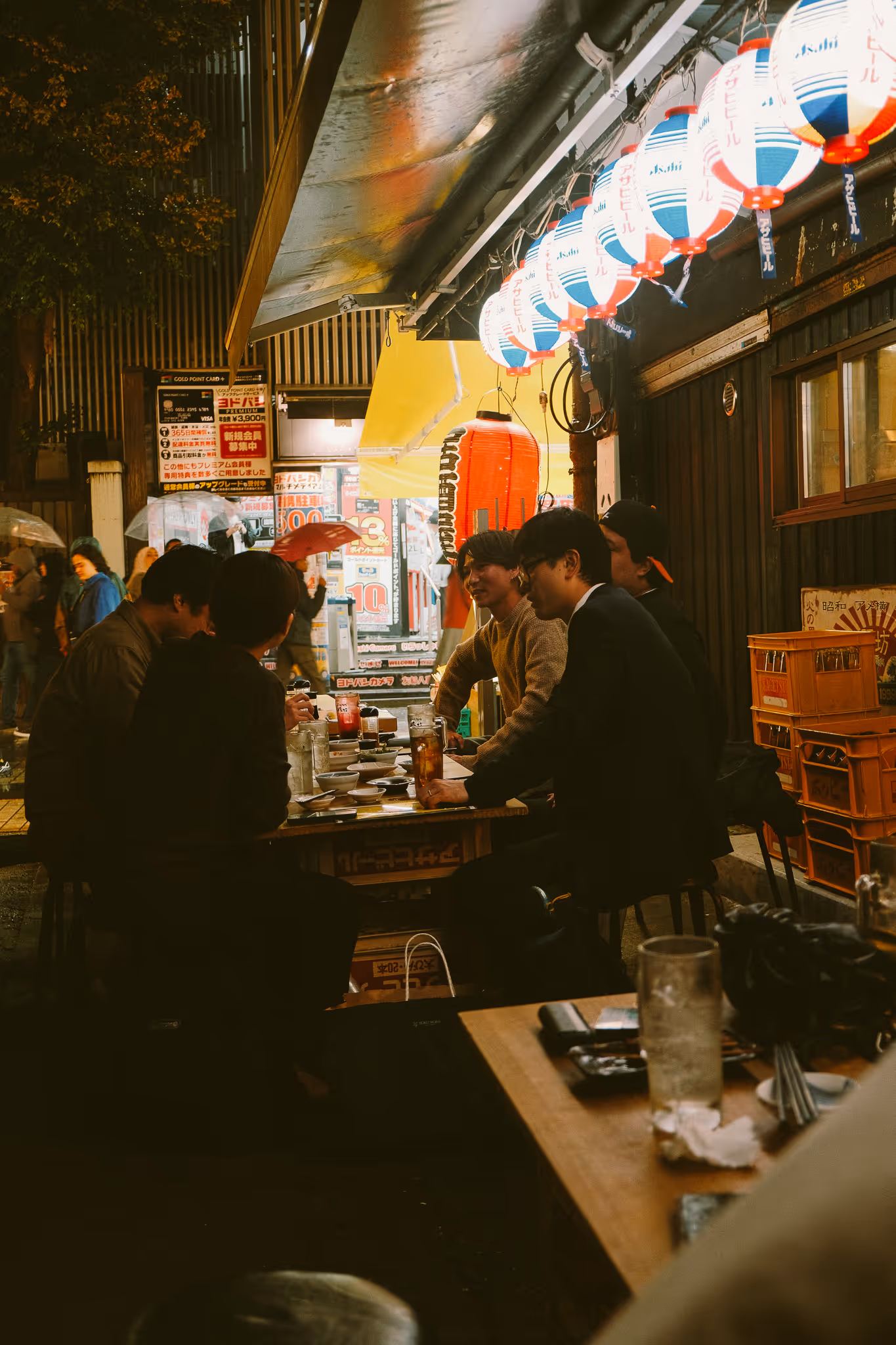 Group of people sitting and chatting at an outdoor Japanese izakaya under hanging red and white lanterns during the evening.