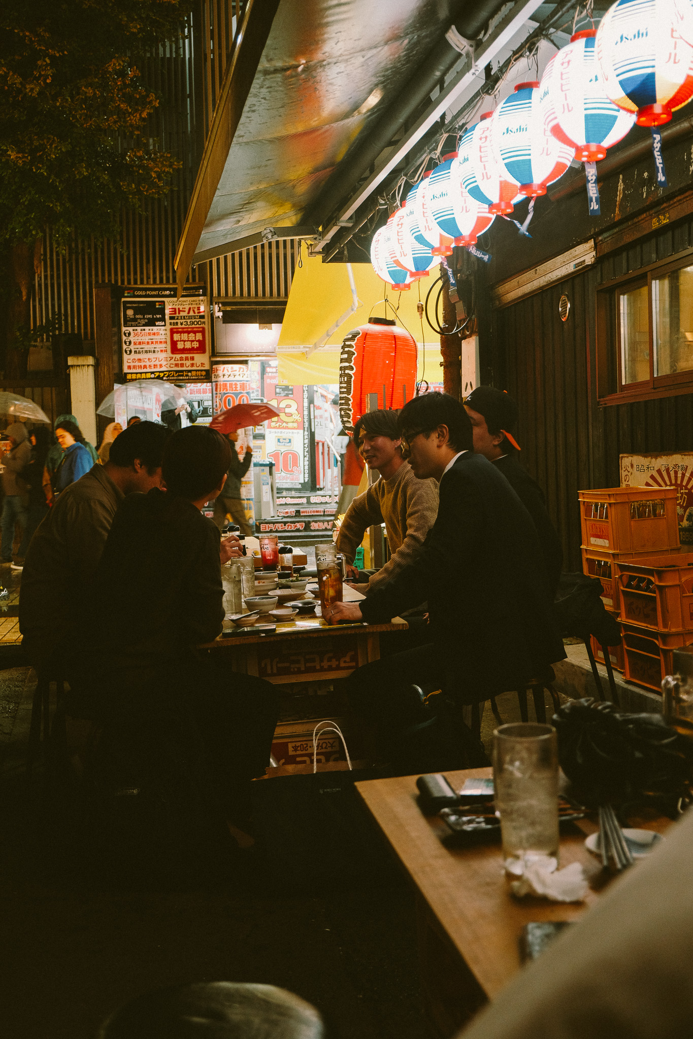 Group of people sitting and chatting at an outdoor Japanese izakaya under hanging red and white lanterns during the evening.