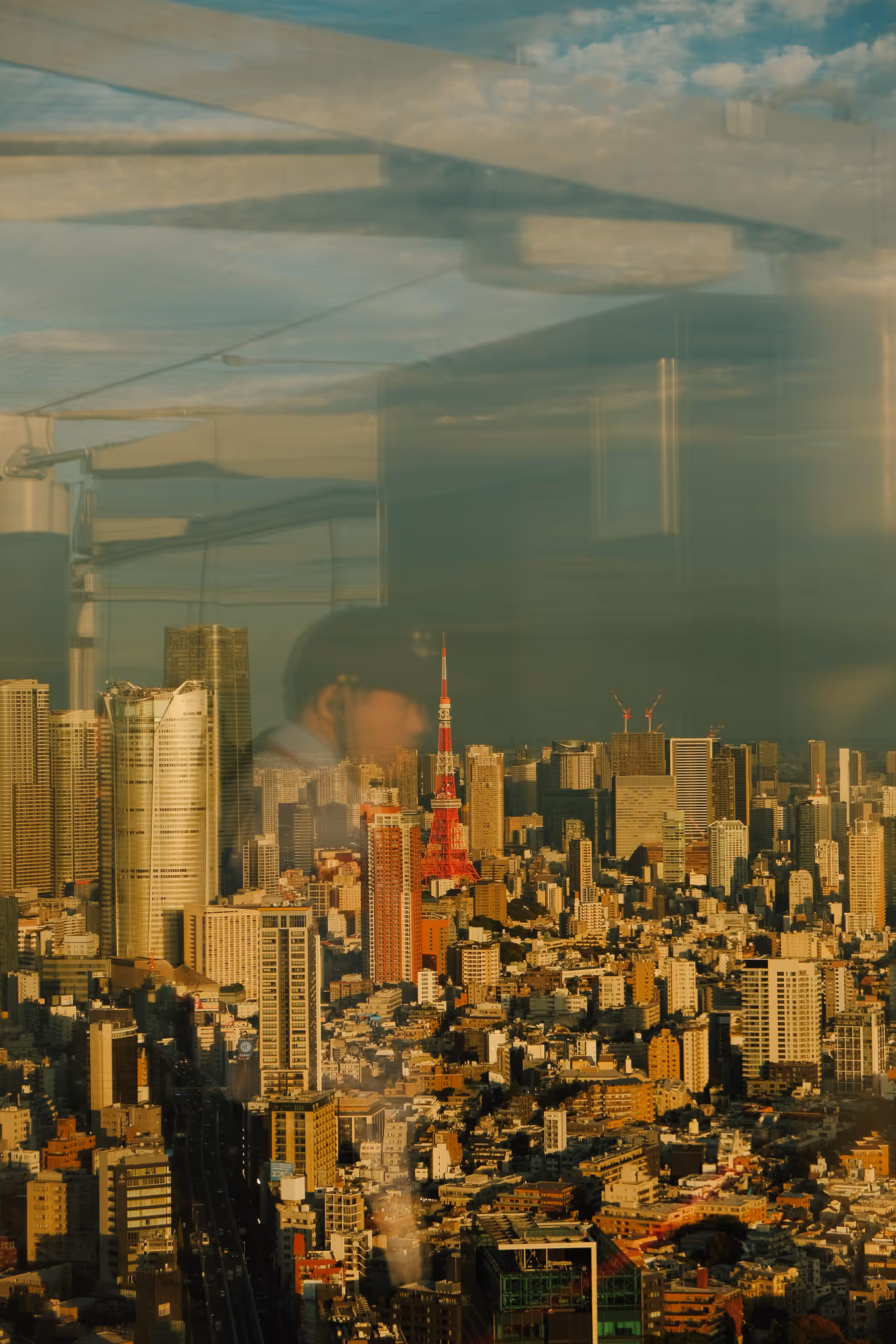 Tokyo cityscape at golden hour with Tokyo Tower prominently in the center and a faint reflection of a man in a cap on the window glass.
