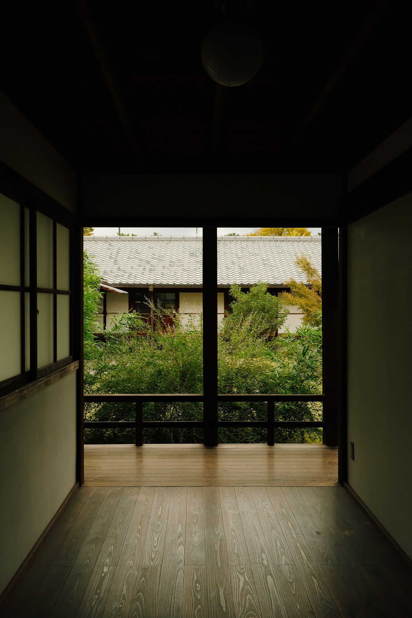 View from a dimly lit wooden hallway looking out onto a porch with greenery and a traditional tiled roof building.