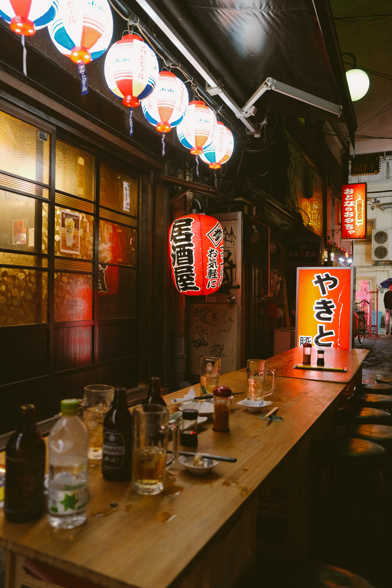 Outdoor nighttime view of a Japanese izakaya with hanging paper lanterns and a wooden table with drinks and condiments.