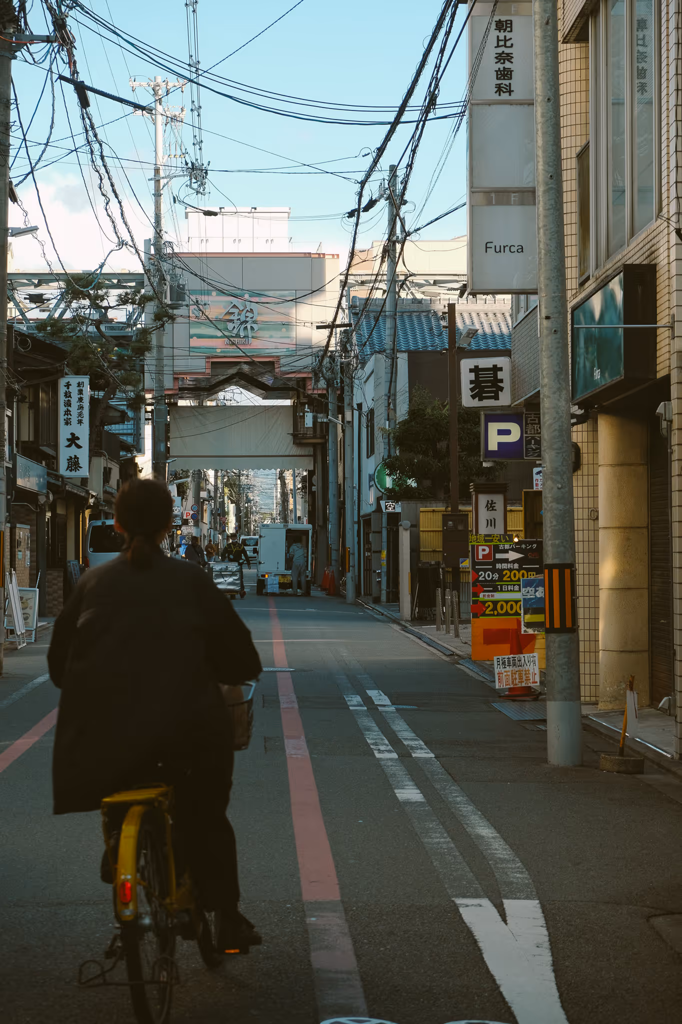 Person riding a yellow bicycle down a narrow urban street lined with buildings and overhead power lines in Japan.
