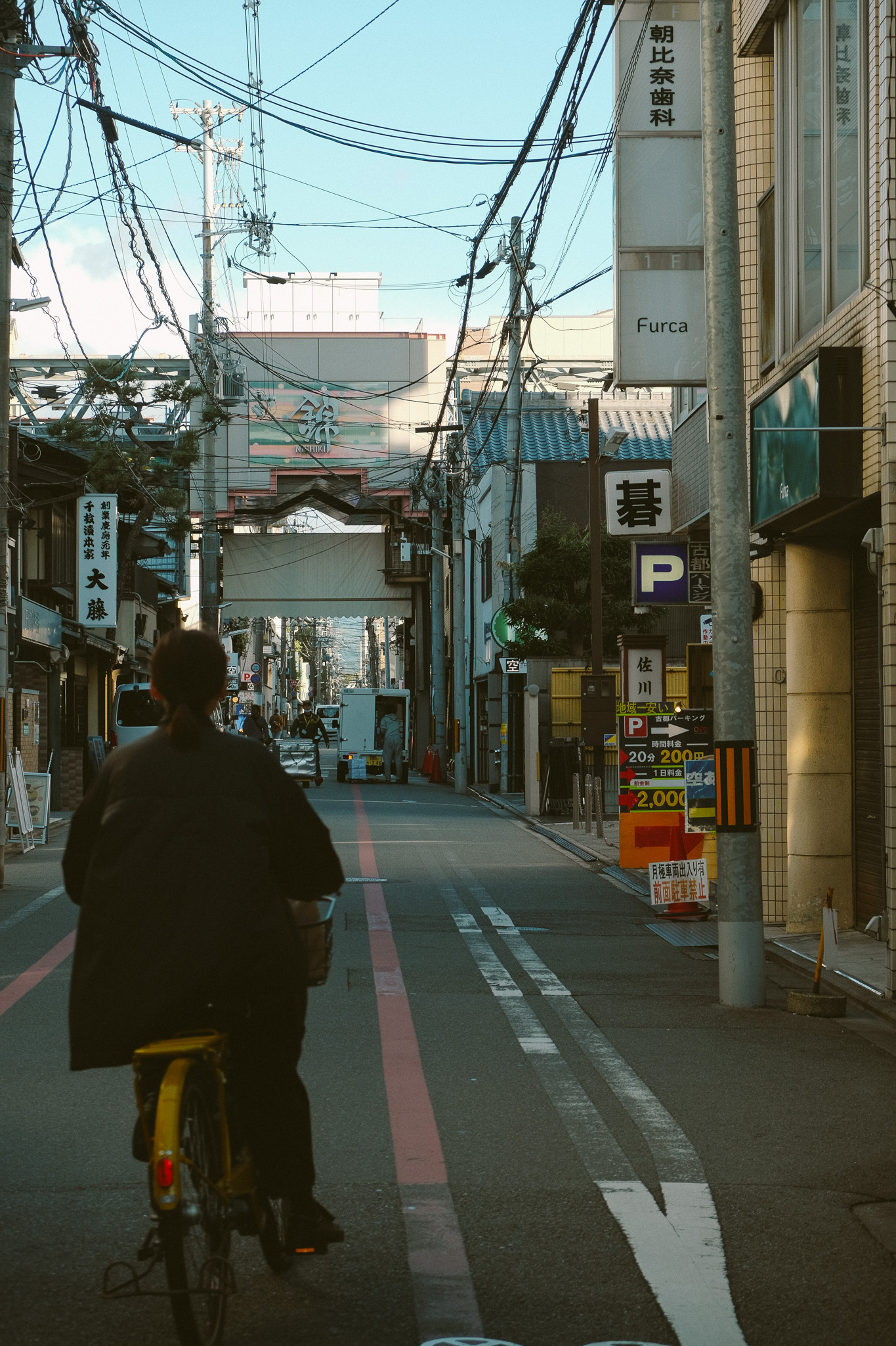 Person riding a yellow bicycle down a narrow urban street lined with buildings and overhead power lines in Japan.