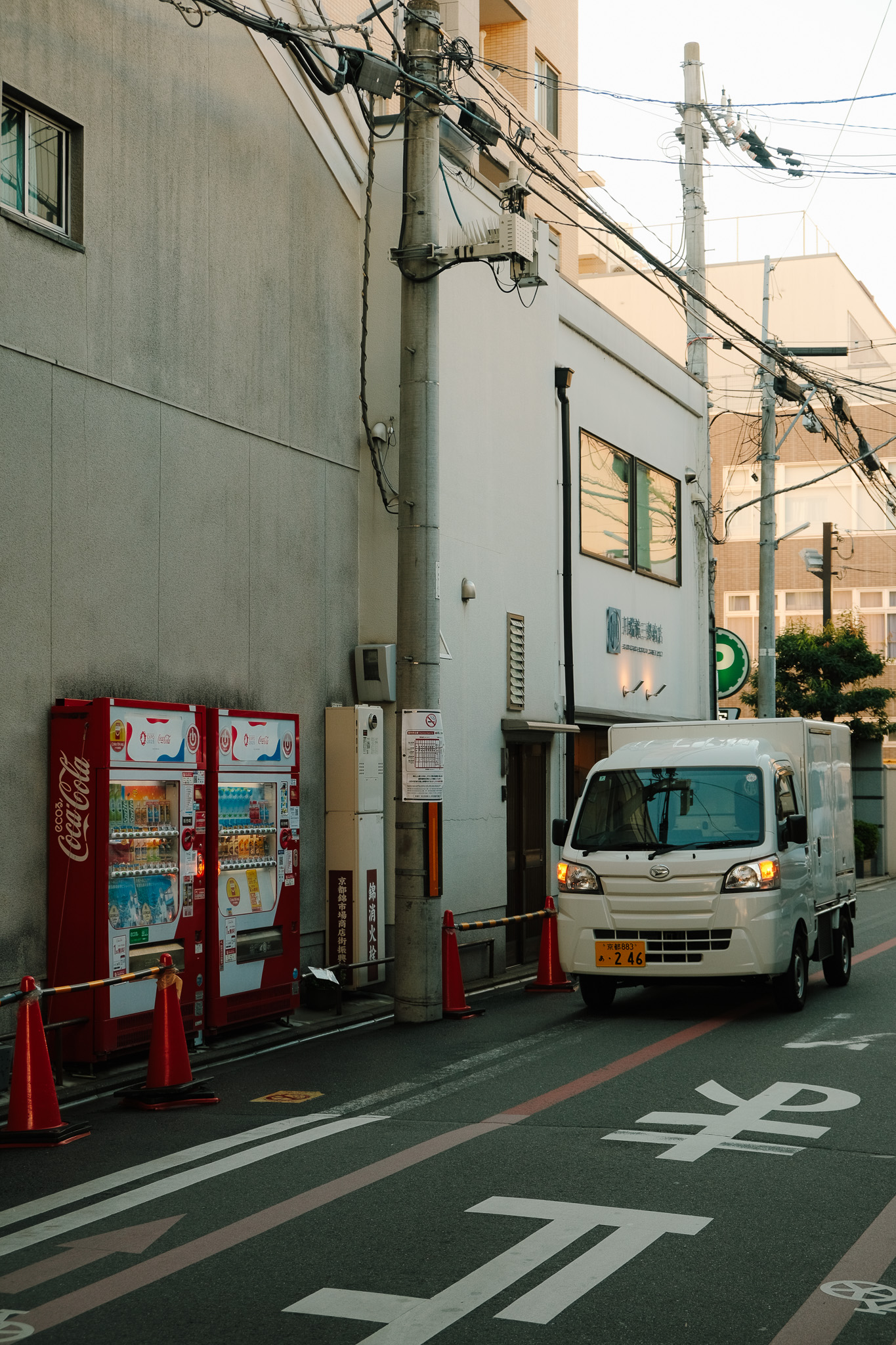 White delivery truck driving on a narrow urban street with red Coca-Cola vending machines and utility poles on the sidewalk.