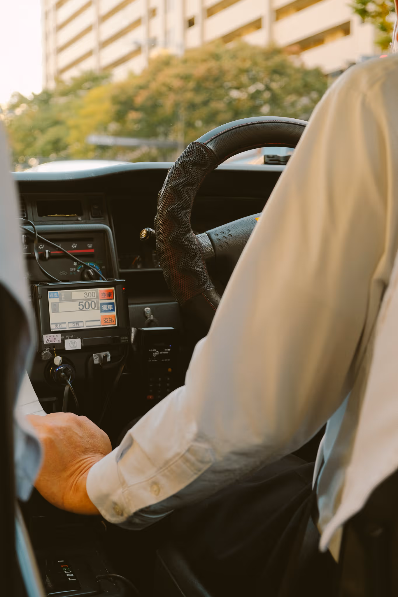 Driver's arm on the steering wheel of a taxi with a fare meter display showing Japanese characters and fare amounts.