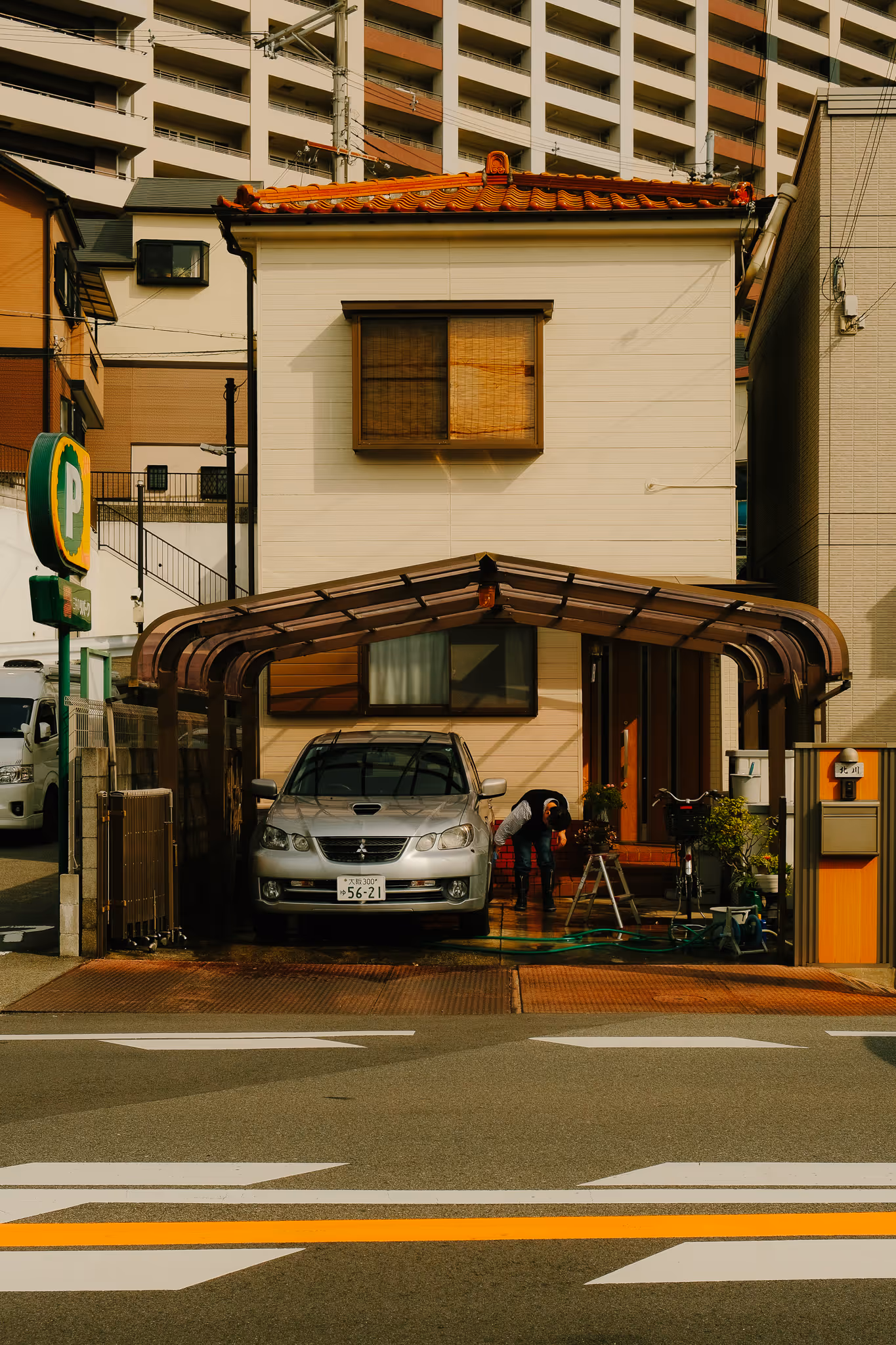 Small beige house with orange tiled roof and carport, a silver car parked inside, and a person bending over near the entrance.
