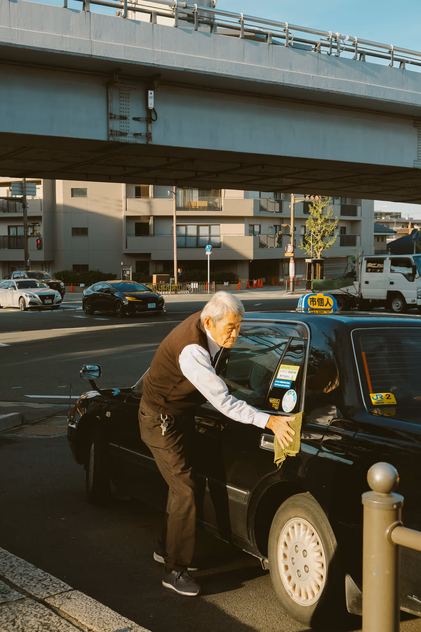 Elderly man cleaning the door of a black taxi parked under an overpass in an urban area.