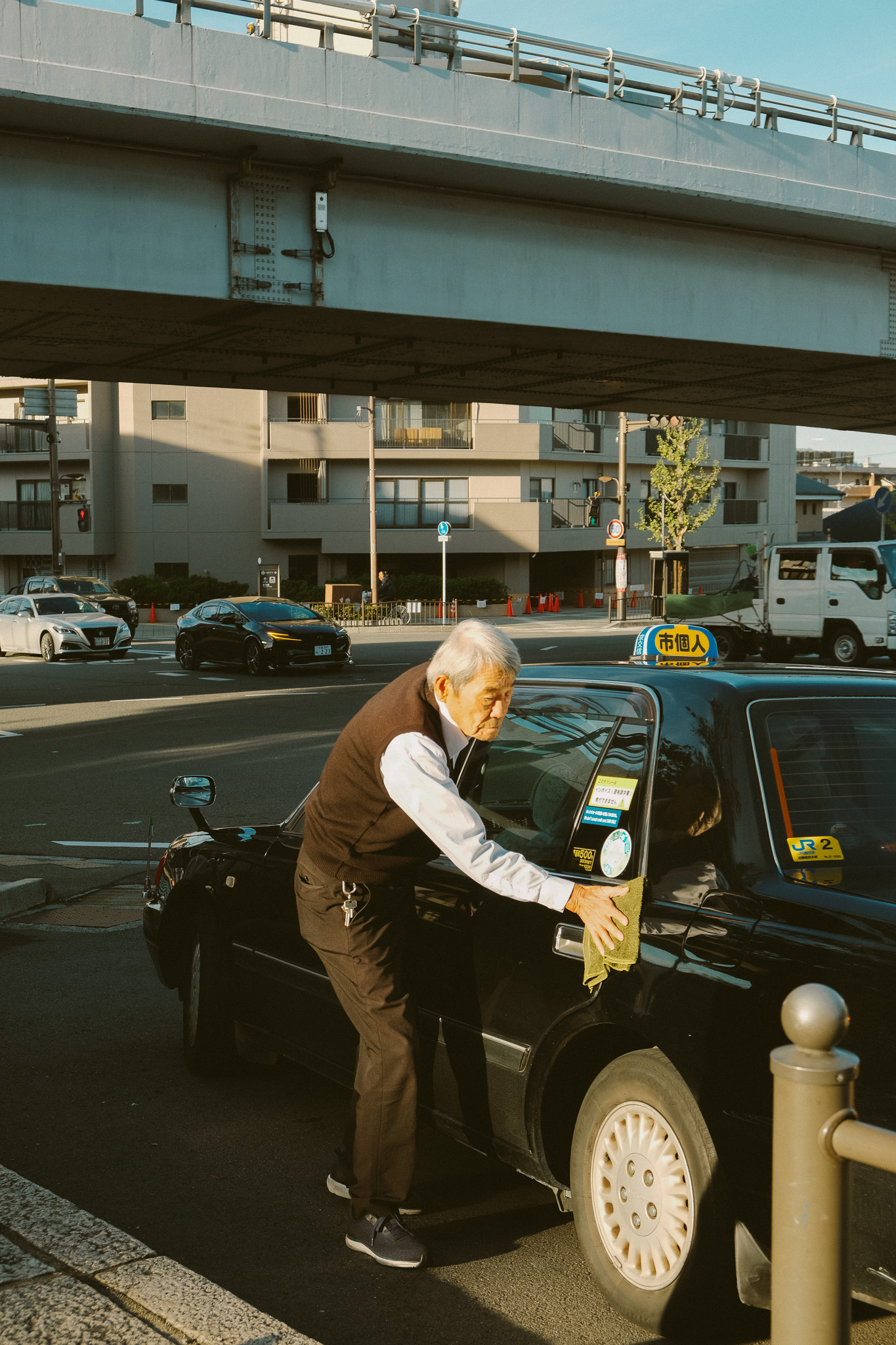 Elderly man cleaning the side of a black taxi under an overpass on a city street.