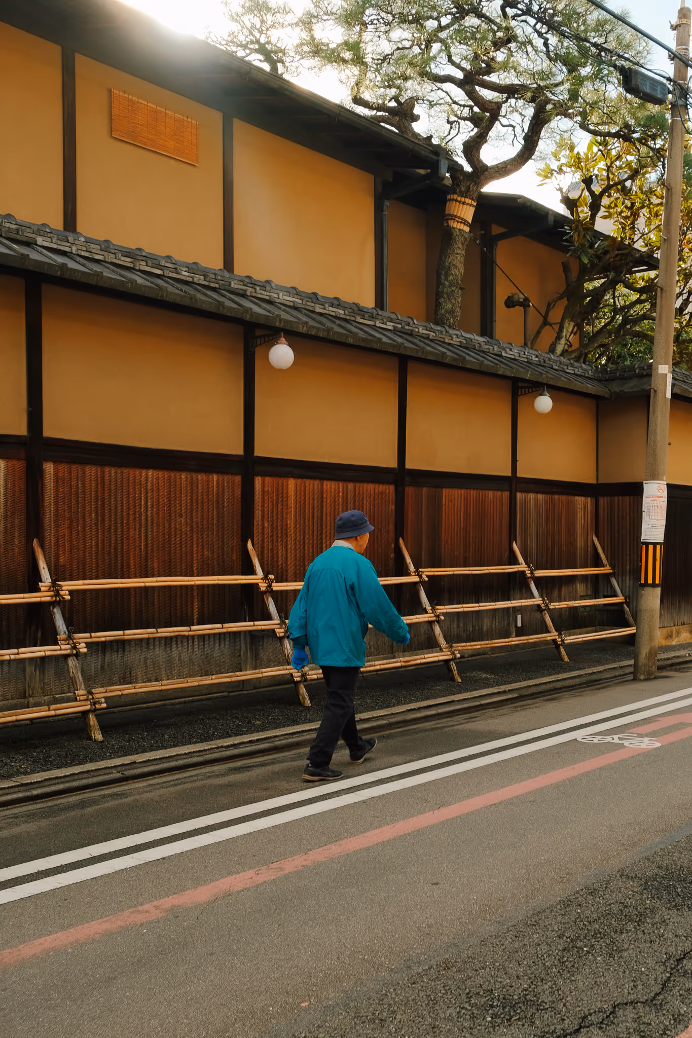 Man in a blue jacket and hat walking along a street beside a traditional Japanese wooden fence and tan building.