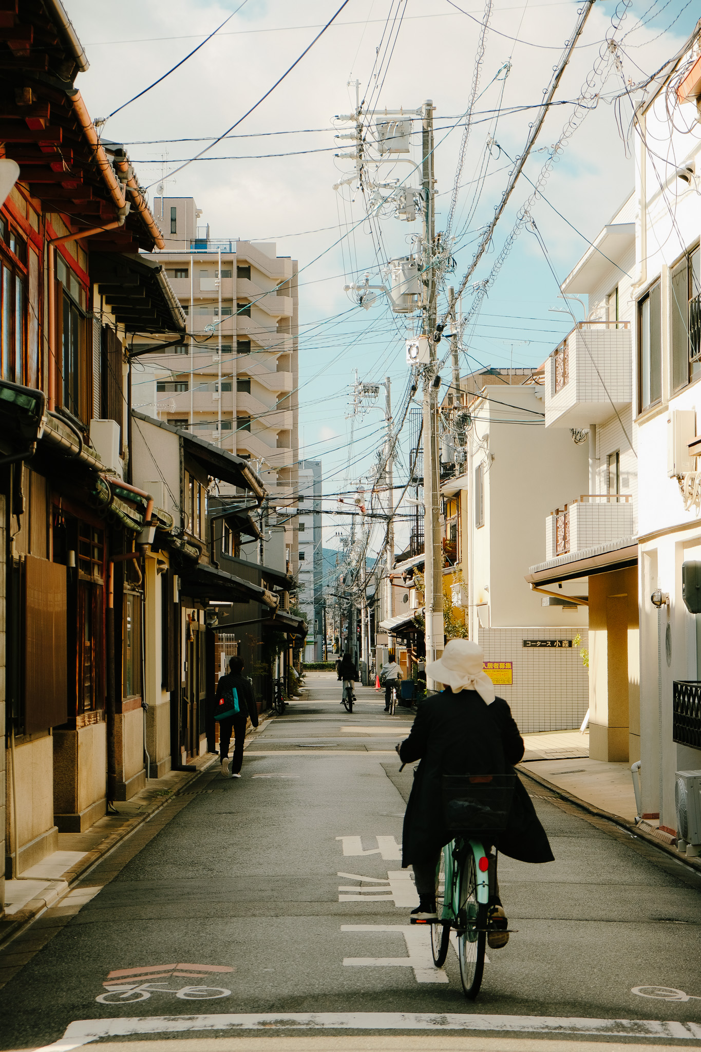Person wearing a hat riding a bicycle down a quiet street lined with buildings and overhead utility wires.