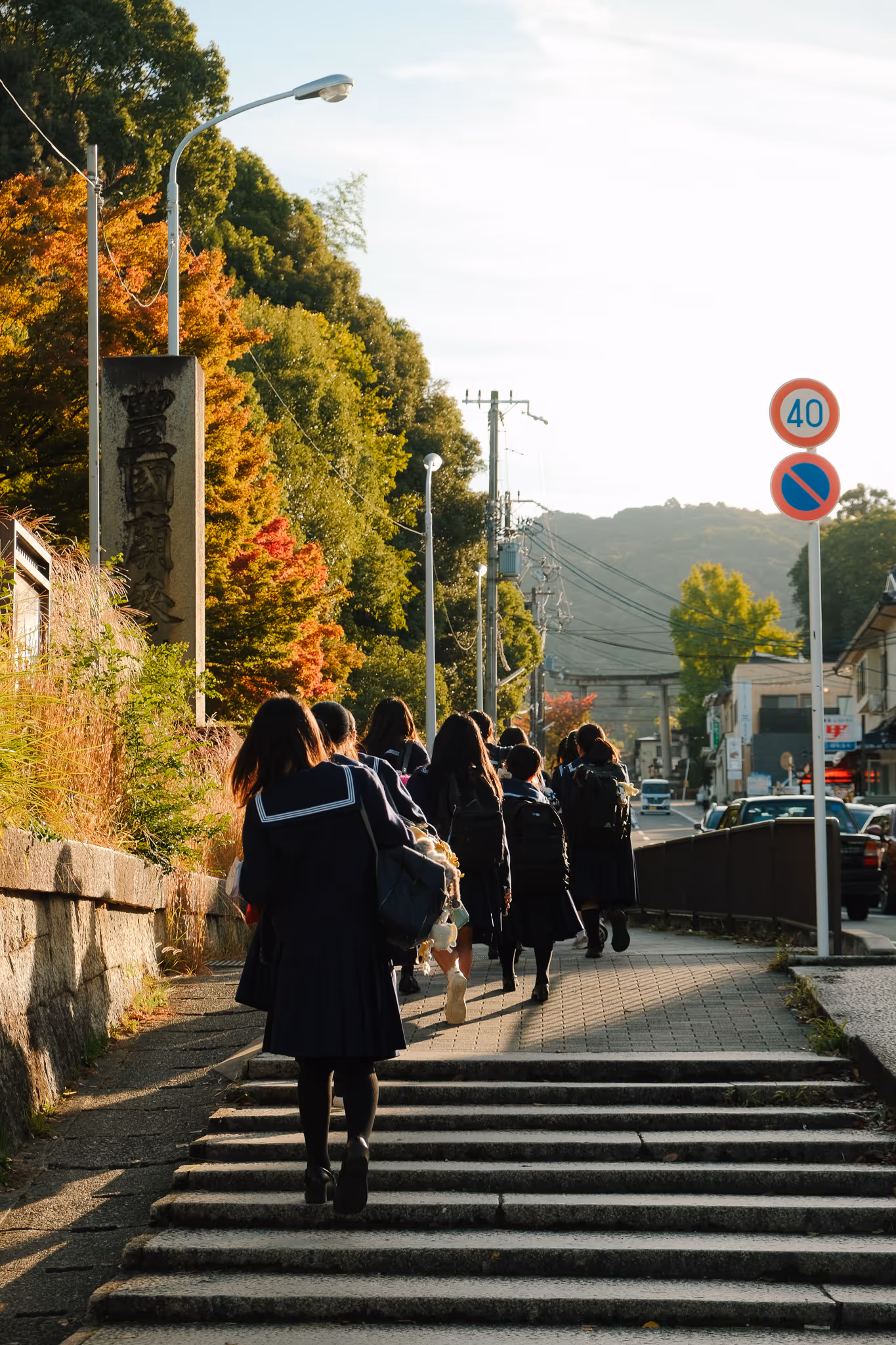 Group of students in navy blue uniforms walking up steps along a sidewalk with autumn trees and traffic signs in a suburban area.