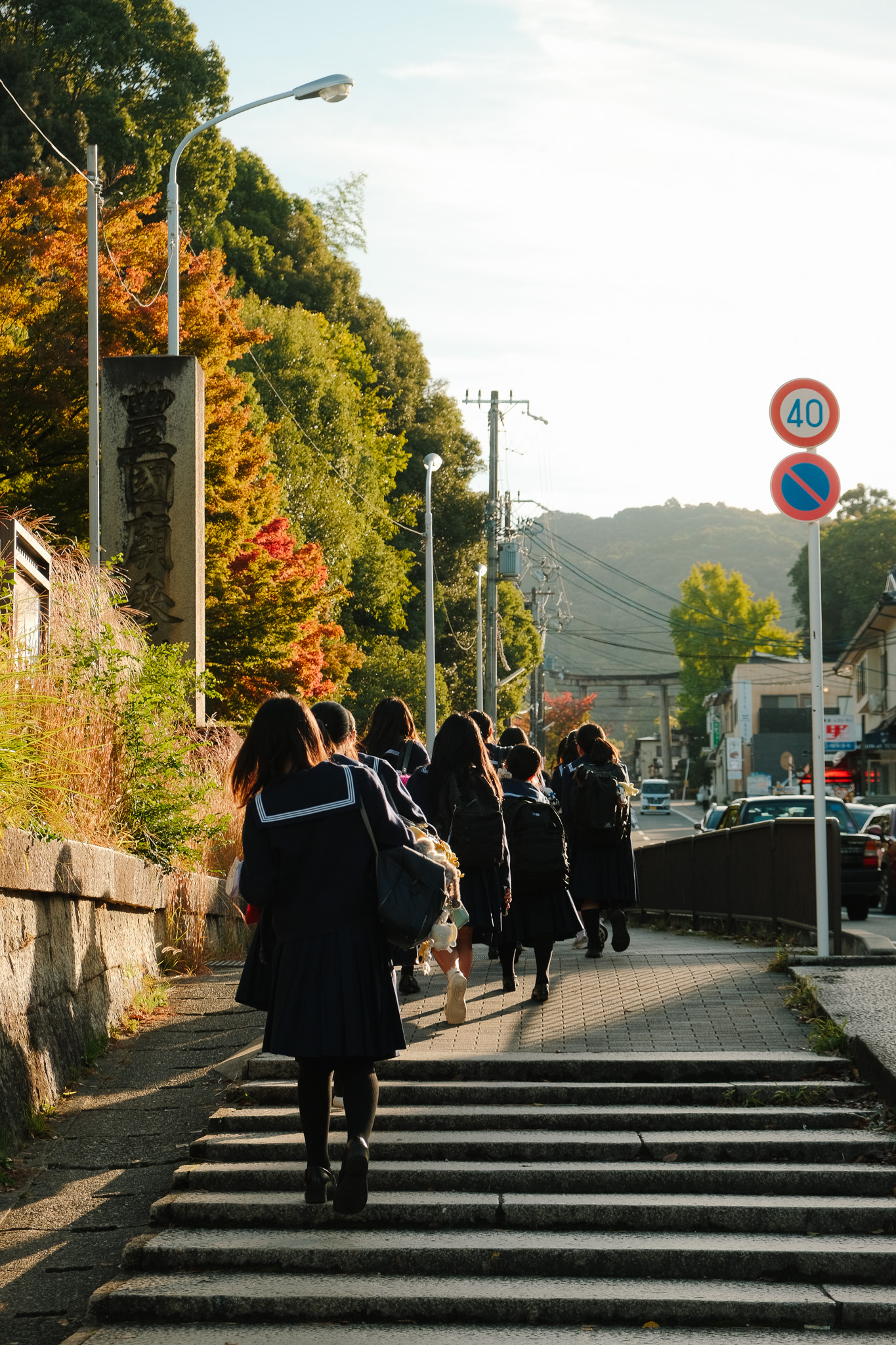 Group of students in navy blue uniforms walking up steps along a sidewalk with autumn trees and traffic signs in a suburban area.