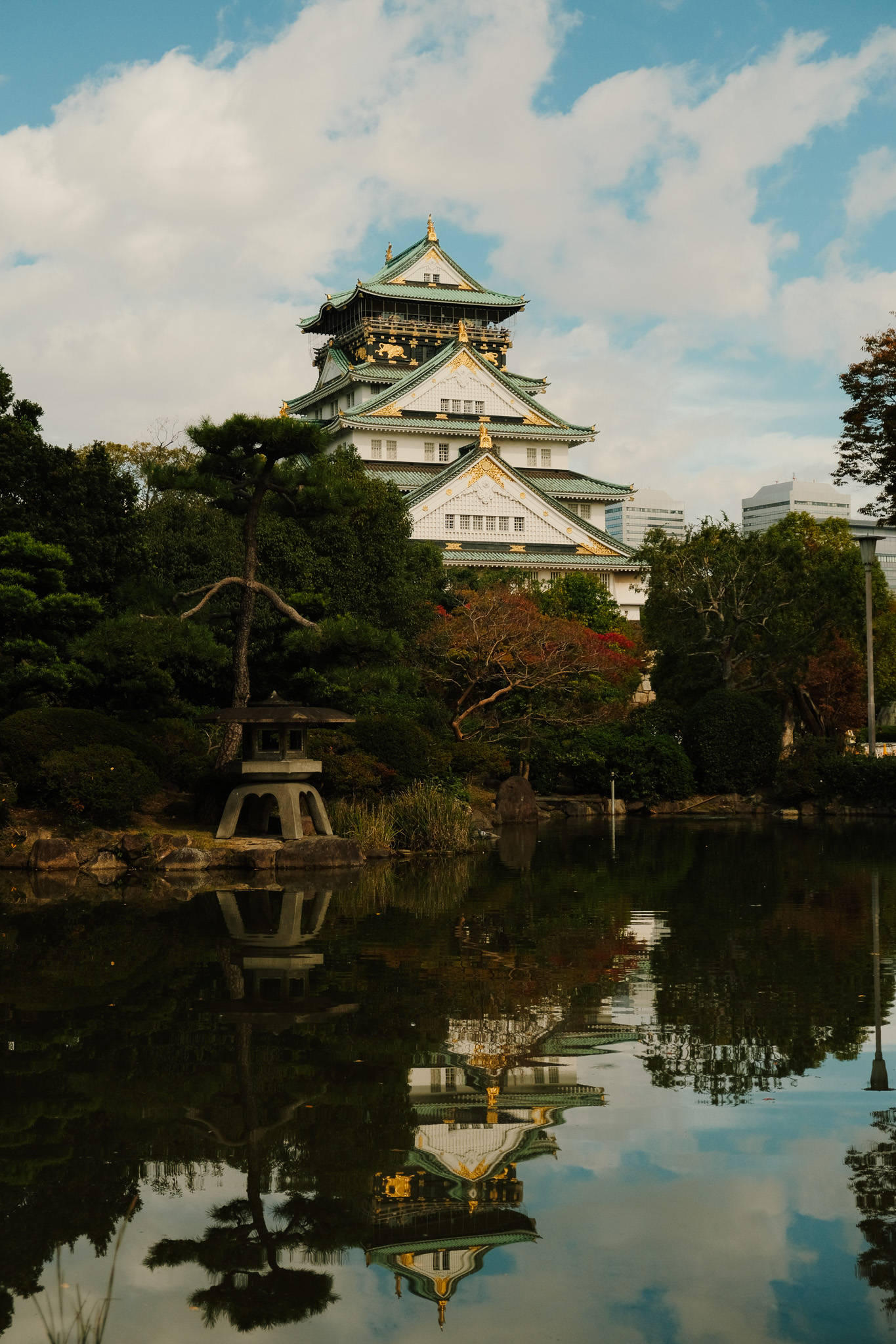 Osaka Castle with green and gold rooftops surrounded by trees and reflected in a calm pond.
