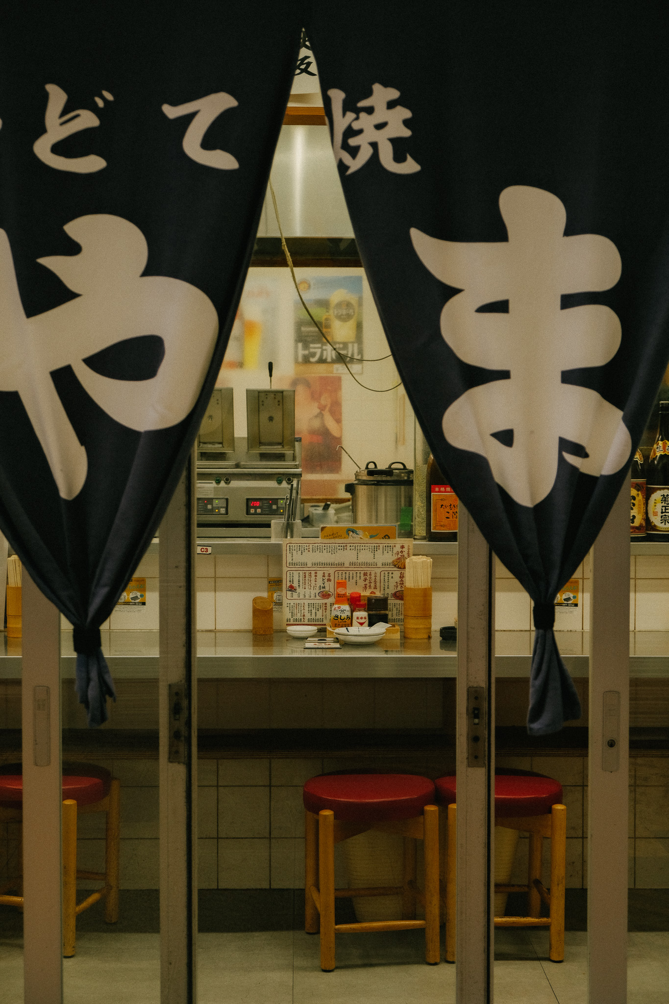 View through a small Japanese restaurant entrance with dark curtains showing a counter, wooden stools, and a menu with condiments.