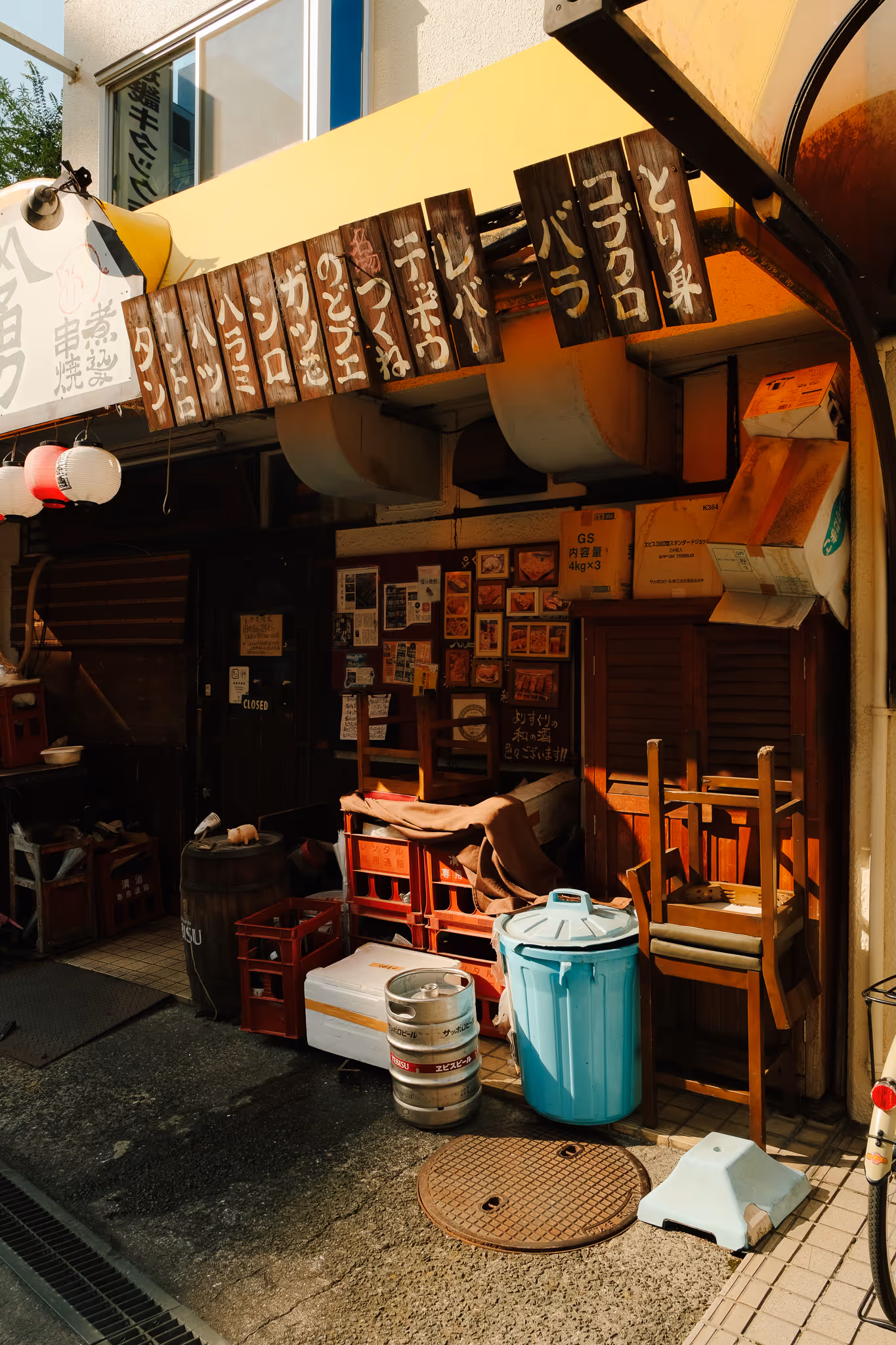 Entrance of a Japanese restaurant with wooden menu boards hanging above, stacked chairs, crates, and a blue trash bin outside.
