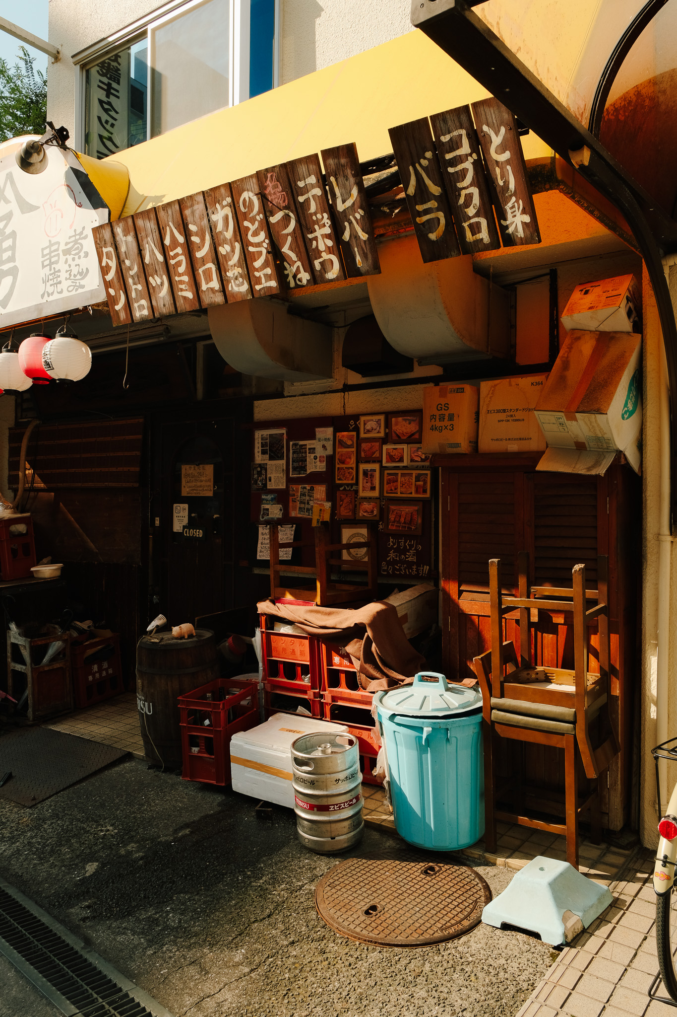 Entrance of a Japanese restaurant with wooden menu boards hanging above, stacked chairs, crates, and a blue trash bin outside.