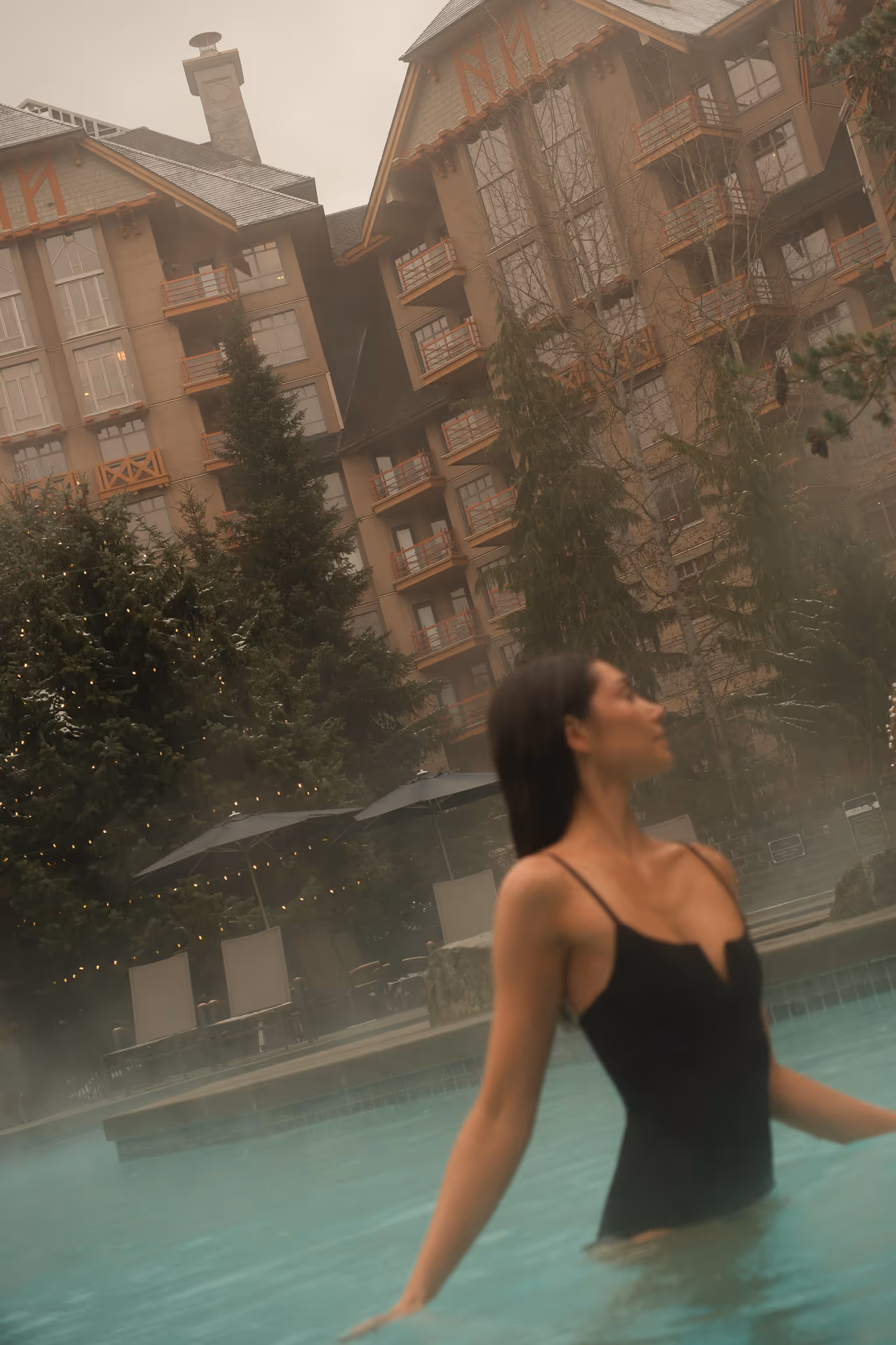 Woman in a black swimsuit standing in a steaming outdoor pool with a large lodge and pine trees in the background.